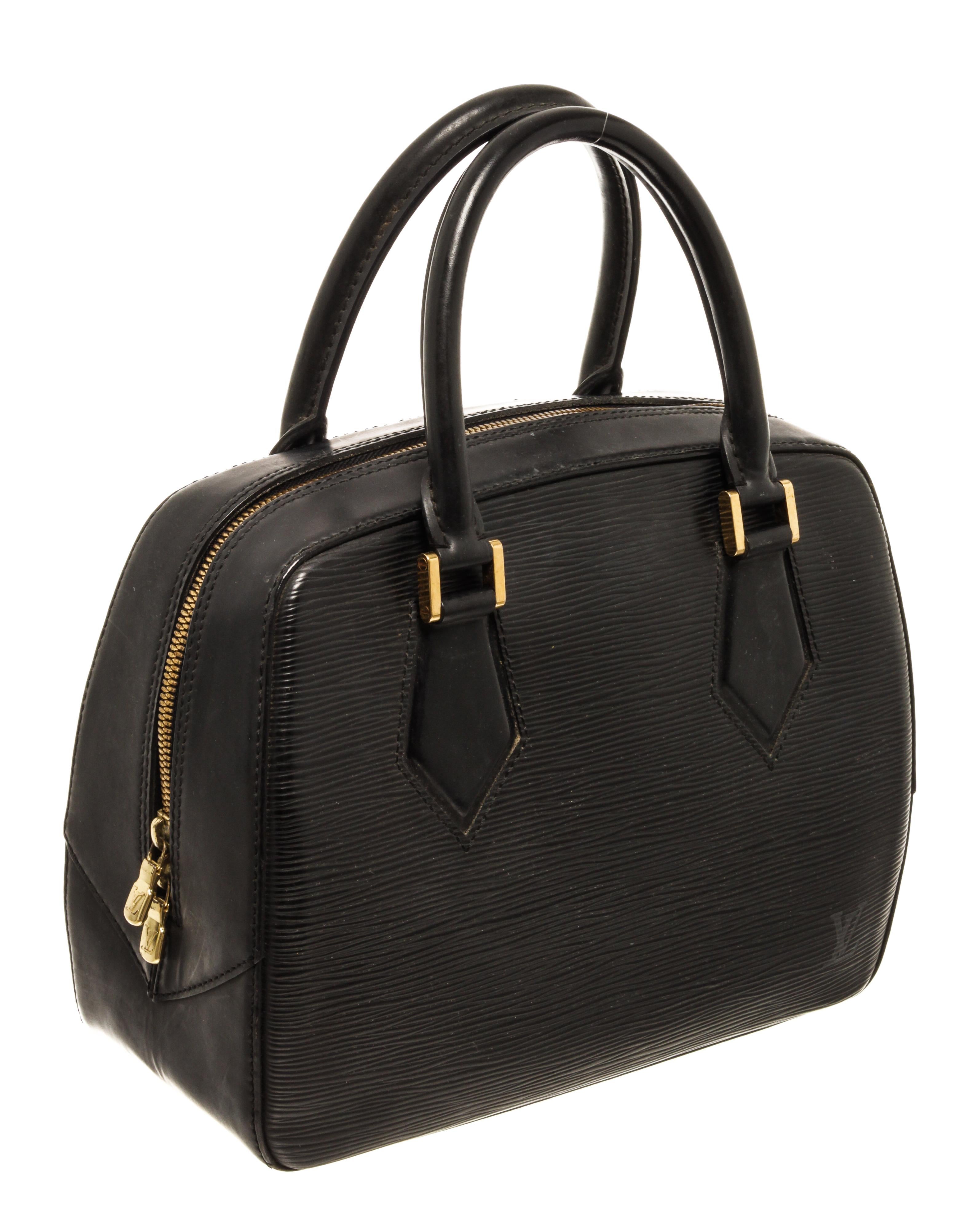 This sophisticated tote is finely crafted of Louis Vuitton signature textured black epi leather. The bag features thin and sturdy rolled leather top handles with squared brass links, and a wrap around zipper. This handbag opens to a alcantara