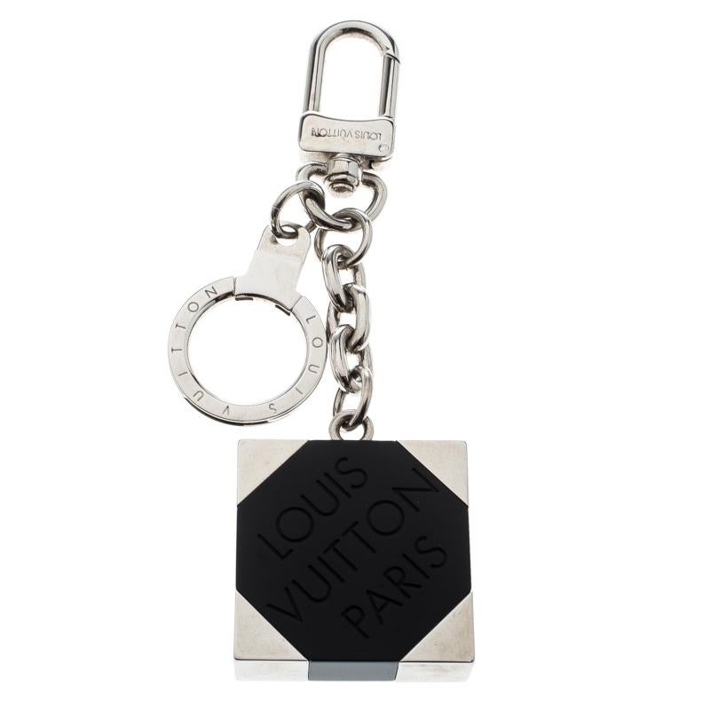 This Louis Vuitton key ring is designed in a silver-tone body featuring a square shaped motif detailed with a black resin panel engraved with the brand detail. The key ring is suspended from a chain which is finished with a clasp closure. This