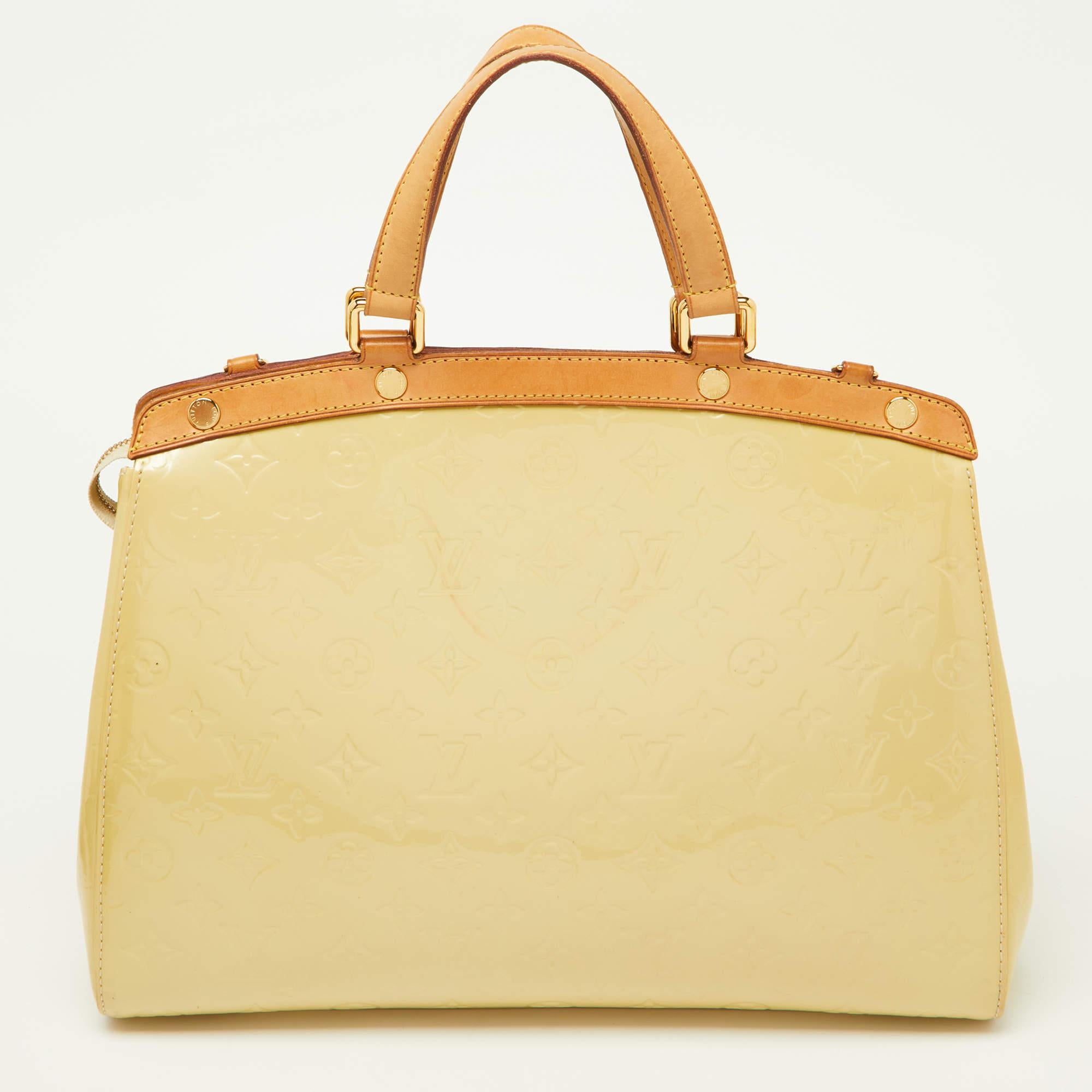 This Brea GM bag from the House of Louis Vuitton will sway you with its precise shape, style, and design. It is made from Blanc Corail Monogram Vernis into a structured, neat silhouette. It flaunts gold-toned fittings, dual top handles, and a