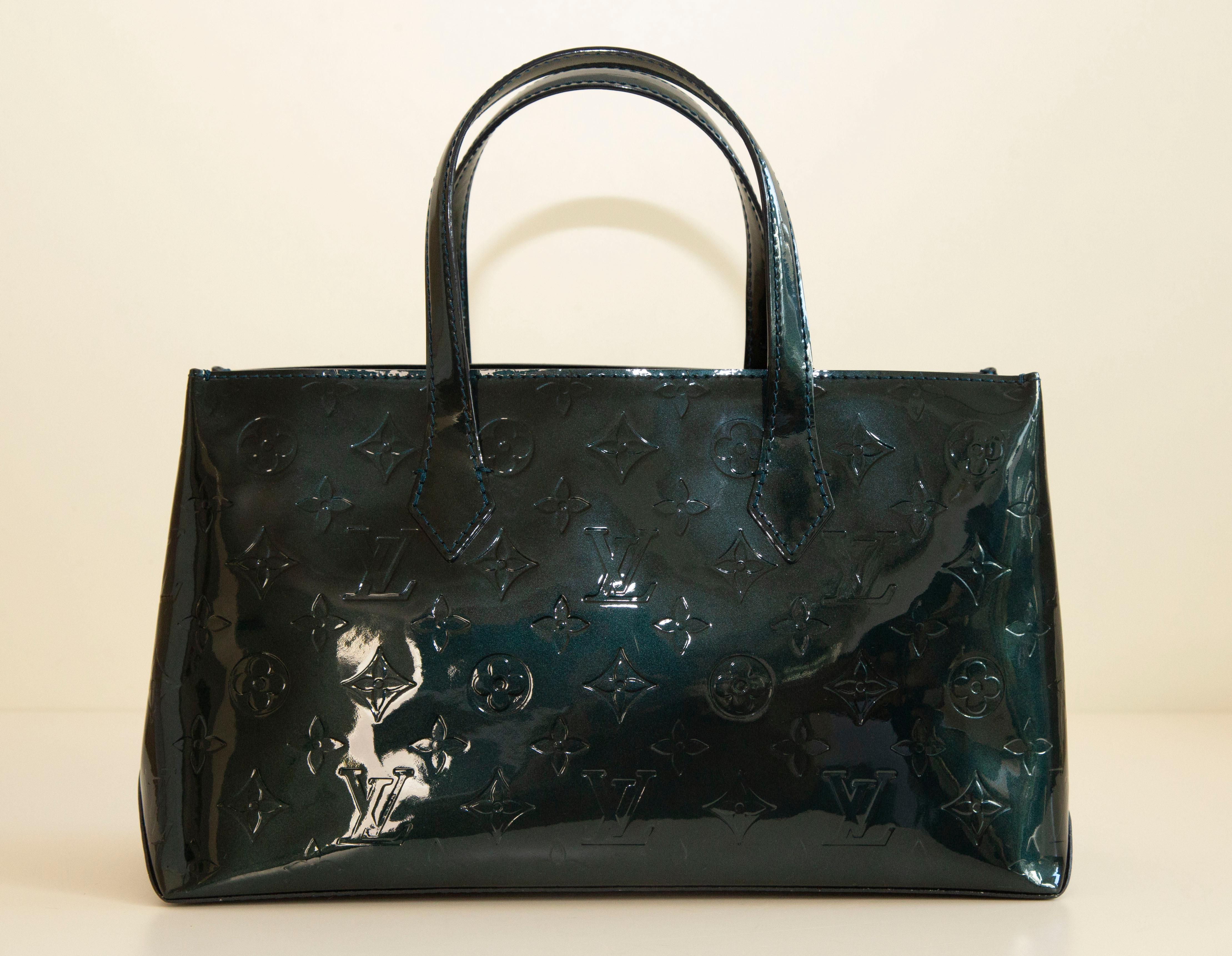 A Louis Vuitton Wilshire PM tote bag crafted from Bleu Nuit (dark green/blue color) Monogram Vernis leather. The interior is lined with a fabric and next to the major compartment it features one side pocket with a zipper. The bag will be delivered