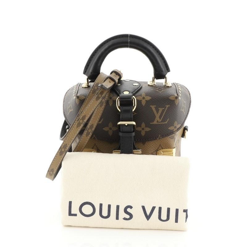 This Louis Vuitton Camera Box Handbag Studded Reverse Monogram Canvas, crafted from brown monogram coated canvas, features leather top handle, stud embellishment, and gold-tone hardware. Its press lock closure opens to a black microfiber interior