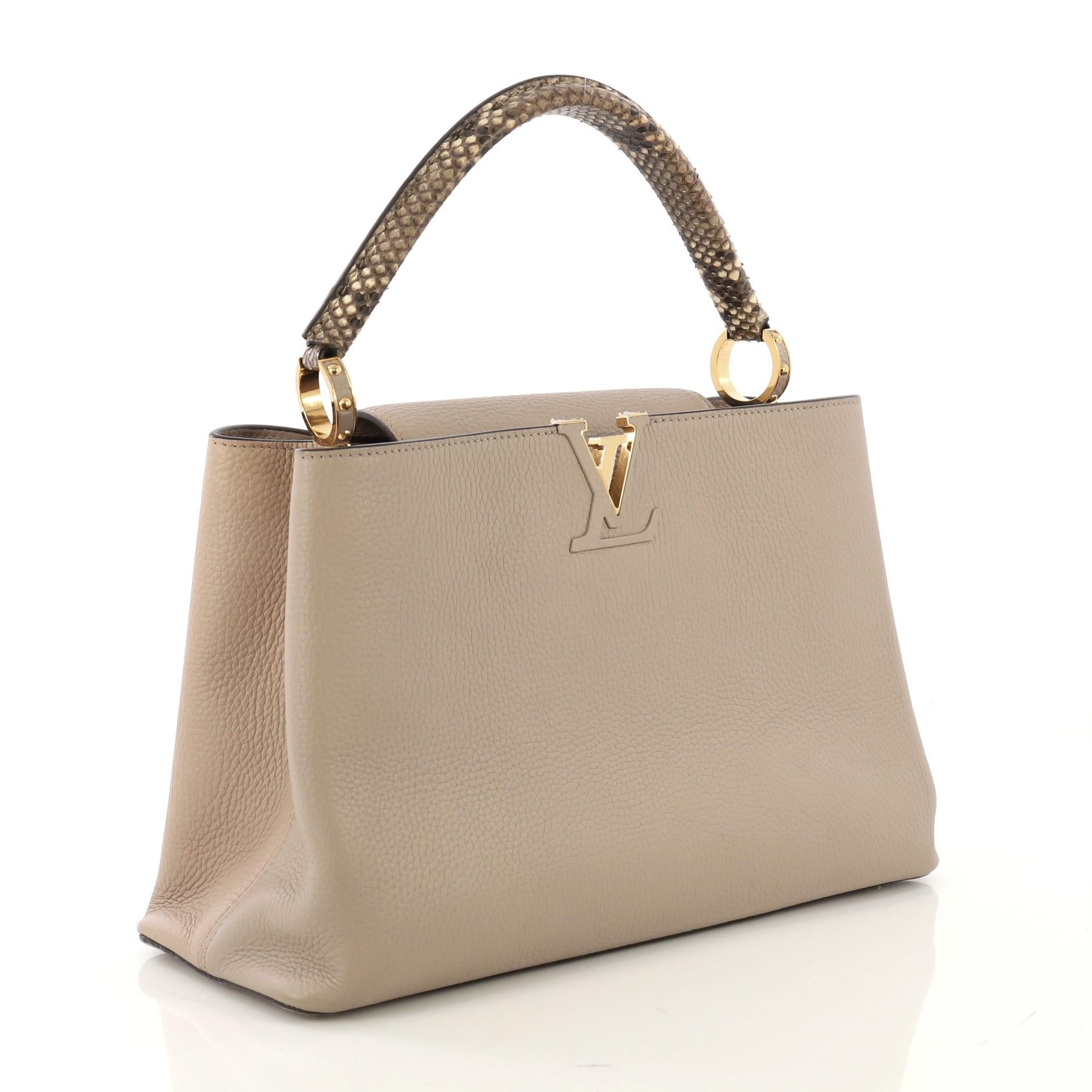 This Louis Vuitton Capucines Handbag Leather and Python MM, crafted in taupe leather and genuine python, features a single rolled genuine python handle secured by jewel-like rings, frontal flap with the classic monogram flower, and gold-tone