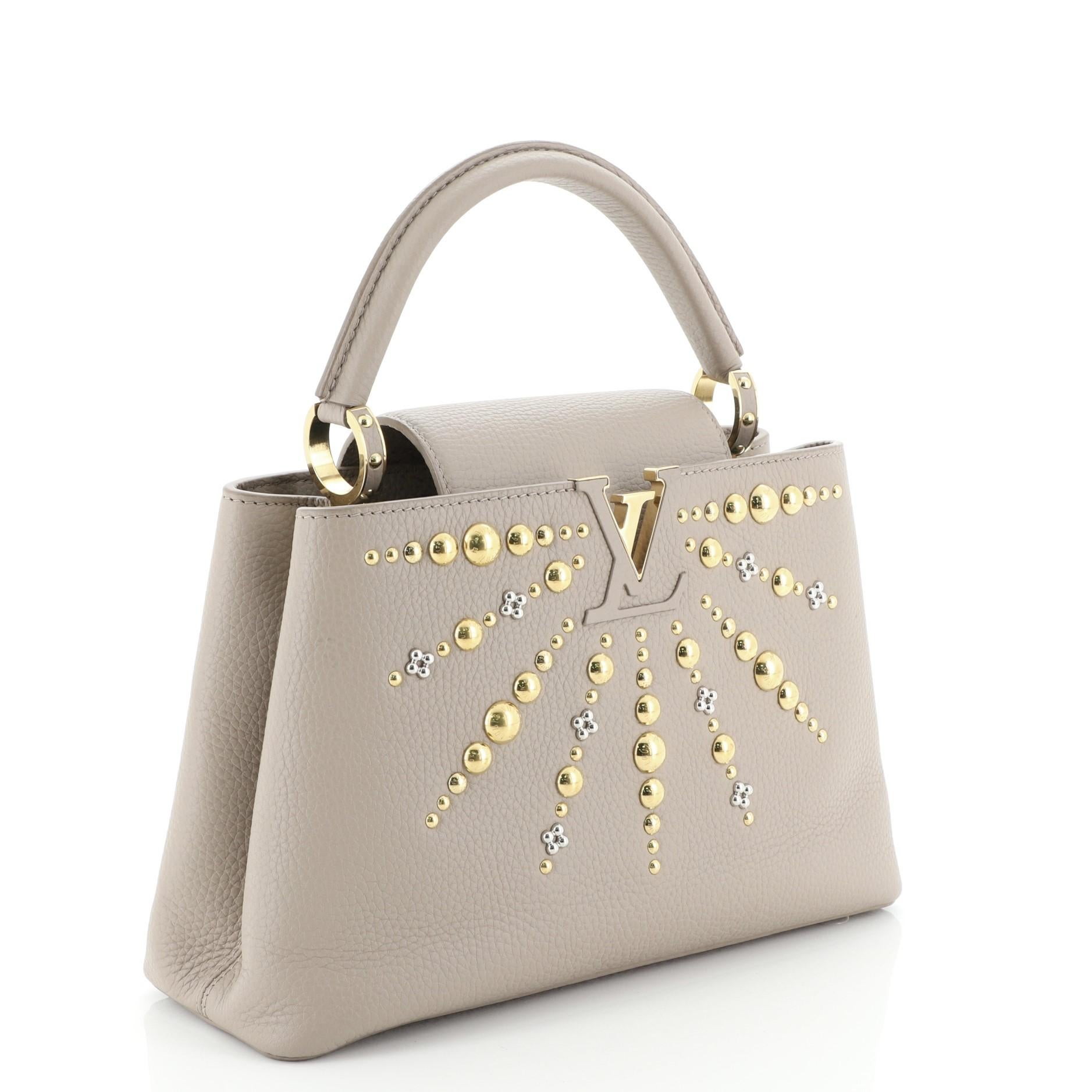 This Louis Vuitton Capucines Handbag Studded Leather PM, crafted in neutral leather, features a single loop leather handle secured by jewel-like rings, stud detailing, frontal flap, and gold and silver-tone hardware. Its flap closure opens to a