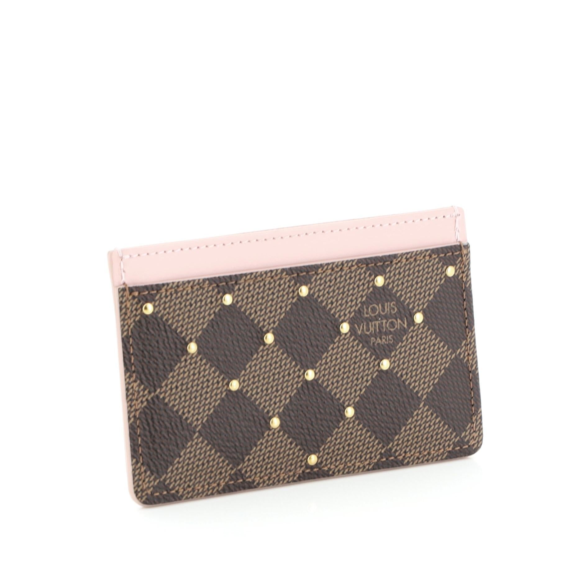 This Louis Vuitton Card Holder Studded Damier, crafted in damier ebene coated canvas, features a leather trim, stud detailing and gold-tone hardware. It opens to a pink leather interior. Authenticity code reads: CA1199. 

Condition: Great. Minor