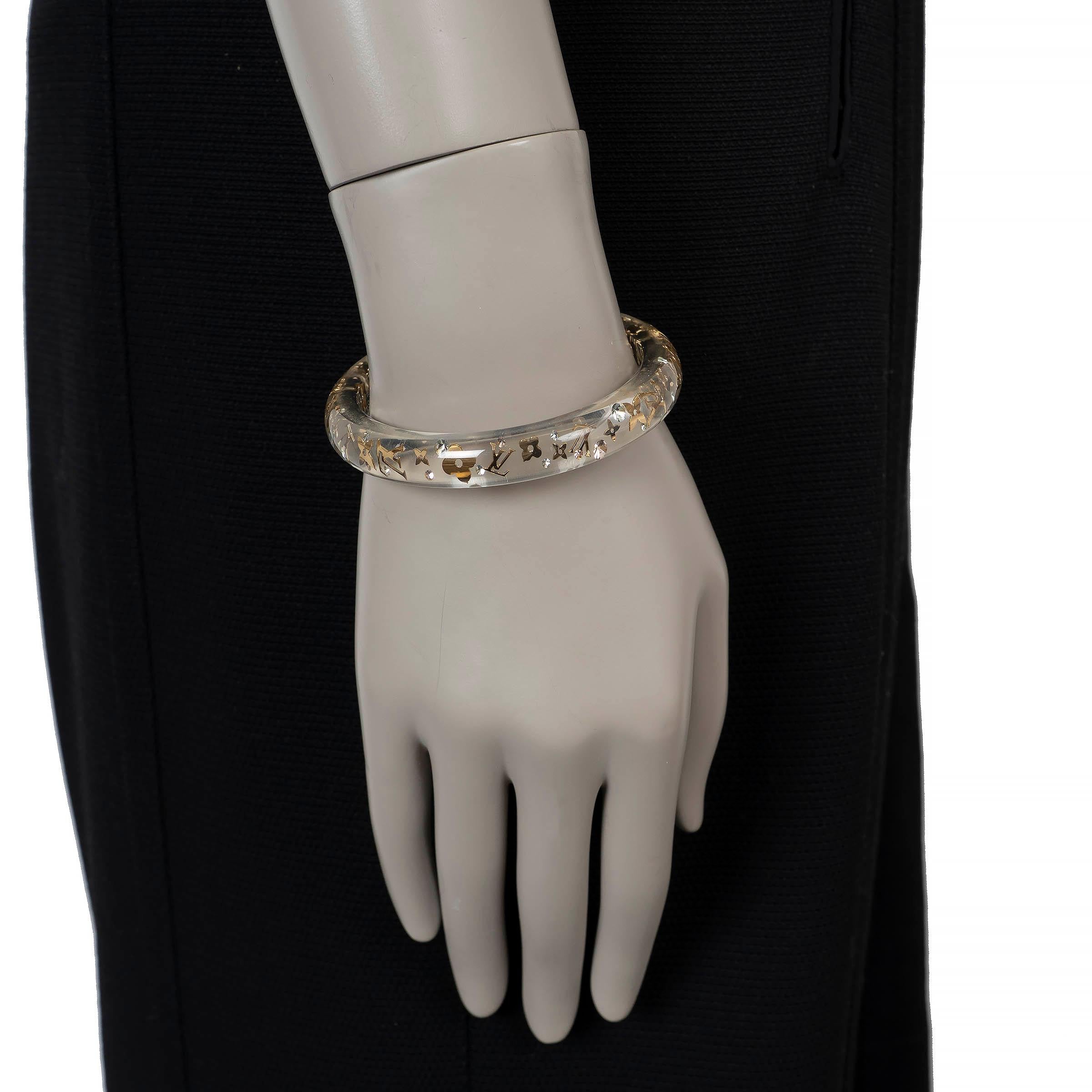 This 100% authentic Louis Vuitton Inclusion bangle bracelet is crafted from clear resin infused with gold-tone Monogram motifs and clear background, creating a striking play of depth and light. The narrow silhouette makes it a bold statement piece,