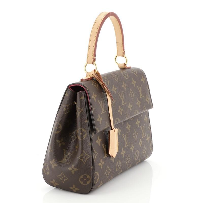 This Louis Vuitton Cluny Top Handle Bag Monogram Canvas BB, crafted in brown monogram coated canvas, features a leather top handle, protective base studs, and gold-tone hardware. Its magnetic closure opens to a pink fabric interior with zip and slip
