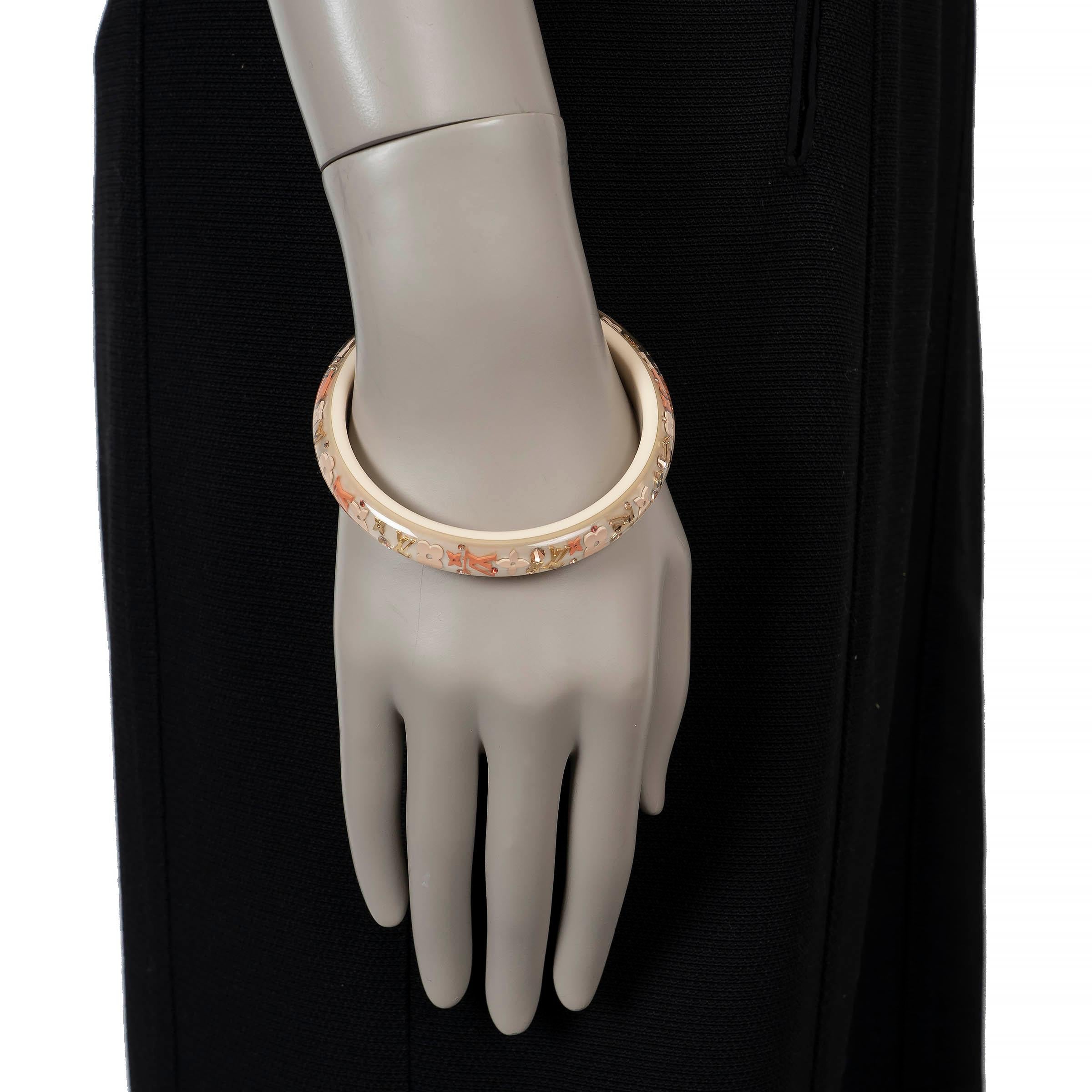 This 100% authentic Louis Vuitton Inclusion bangle bracelet is crafted from clear resin infused with gold-tone Monogram motifs and cream background, creating a striking play of depth and light. The narrow silhouette makes it a bold statement piece,