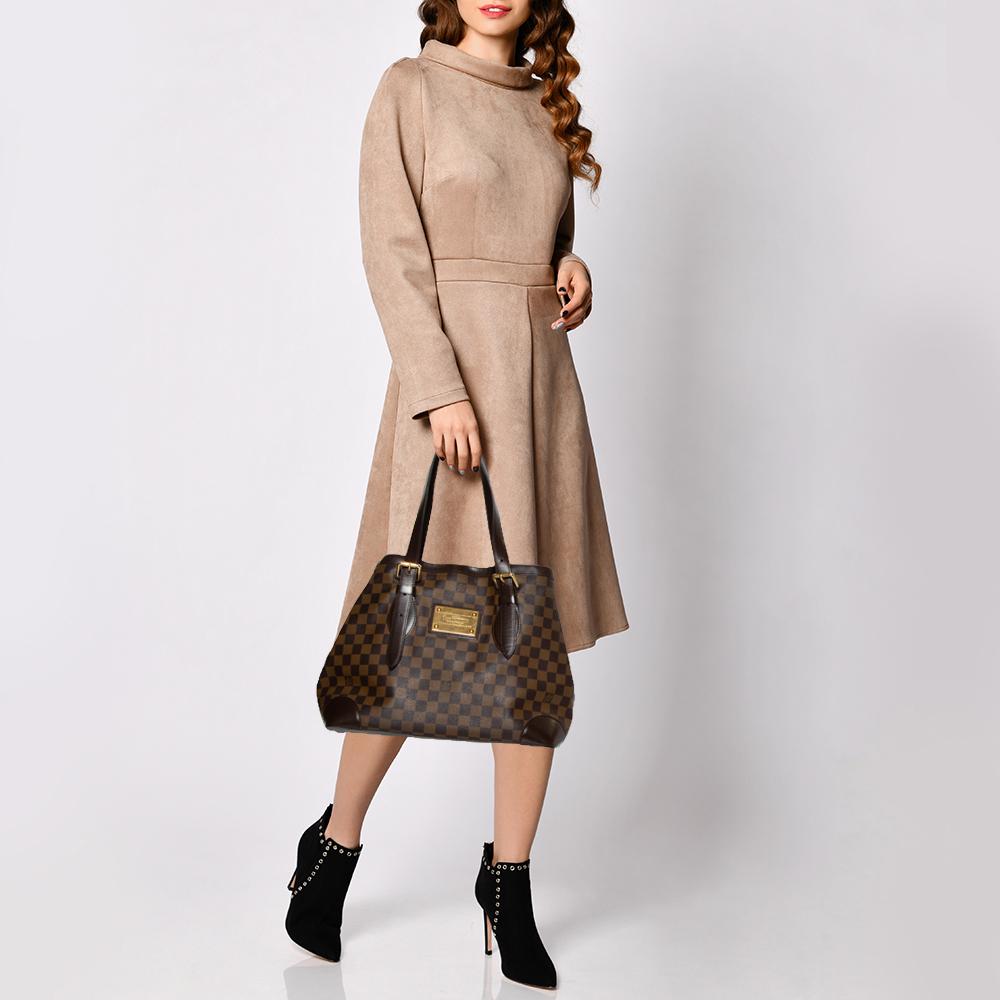 Handbags from Louis Vuitton enjoy widespread popularity owing to their high style and functionality. This Hampstead bag is no exception. Crafted from LV's signature Damier Ebene canvas and leather, the bag comes with two flat top handles and an