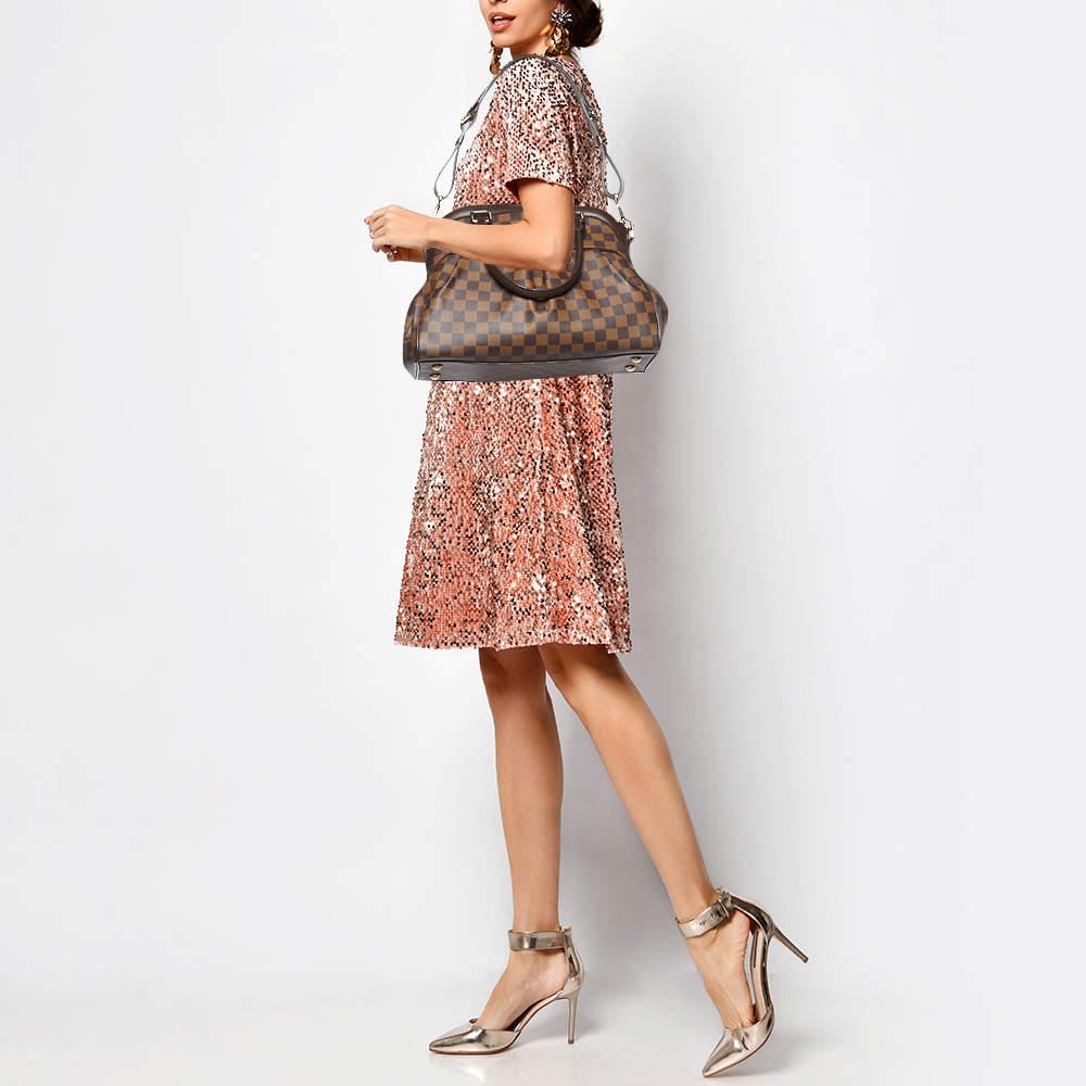 This pre owned Trevi bag by Louis Vuitton is crafted from Damier Ebene canvas and it features two leather handles and an optional bag strap. It has gold tone hardware protective metal feet and a top zip that opens to a spacious Alcantara
