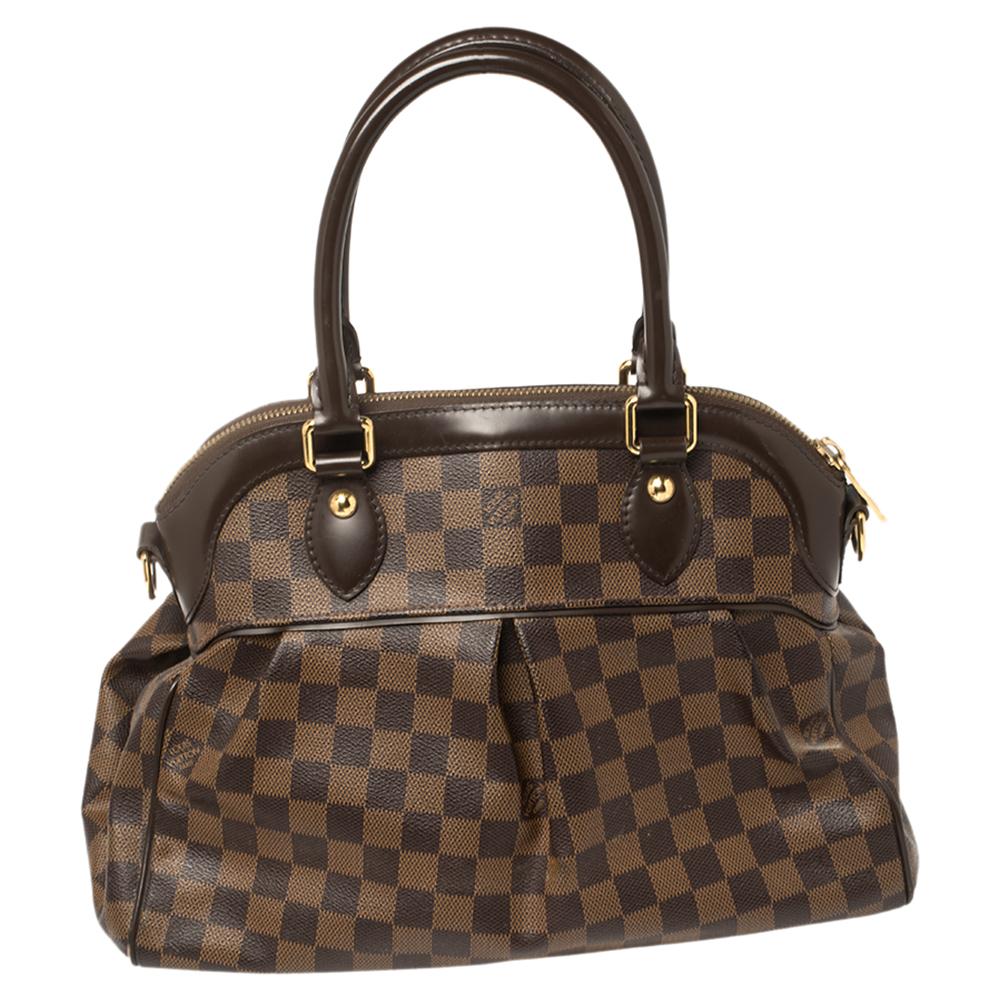 This Trevi bag by Louis Vuitton has been crafted from Damier Ebene coated canvas and it features two leather handles. It has gold-tone hardware, protective metal feet, and a top zip that opens up to a spacious Alcantara compartment. This finely-made