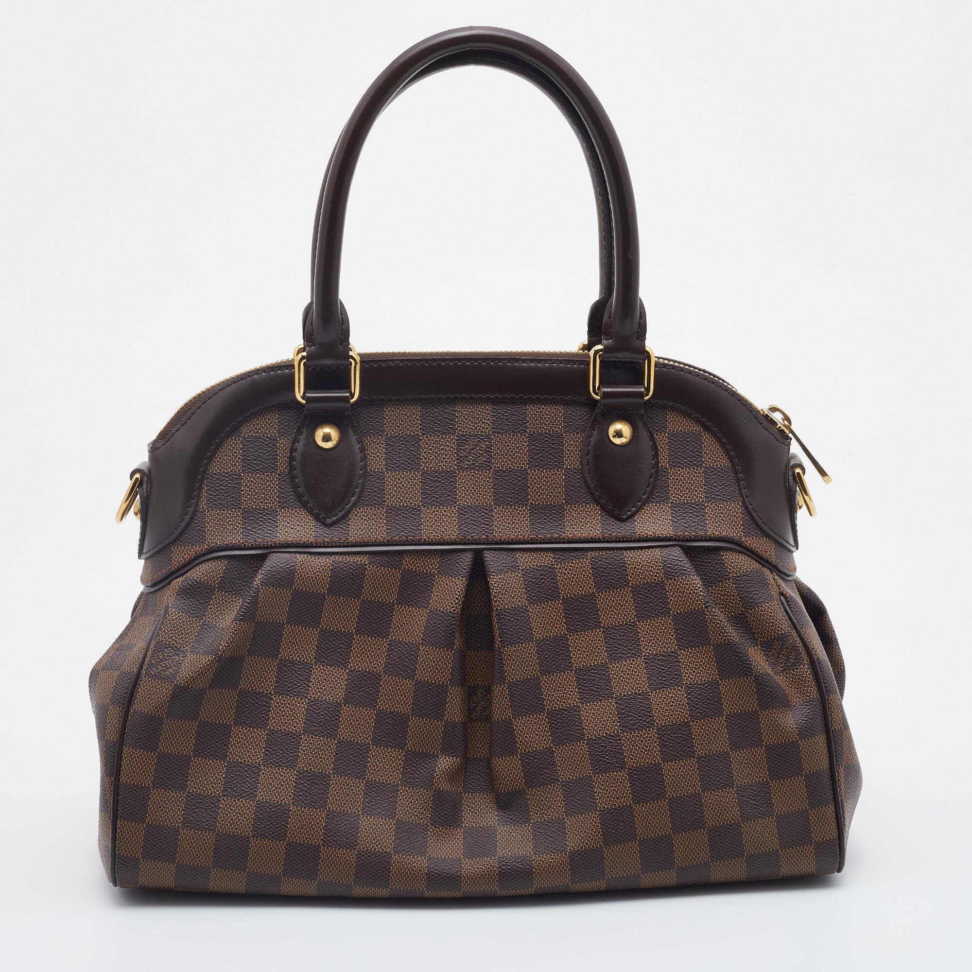 This pre-owned LV Trevi bag is crafted from Damier Ebene canvas, and it features two leather handles and an optional bag strap. It has gold-tone hardware, protective metal feet, and a top zip that opens up to a spacious Alcantara compartment. This
