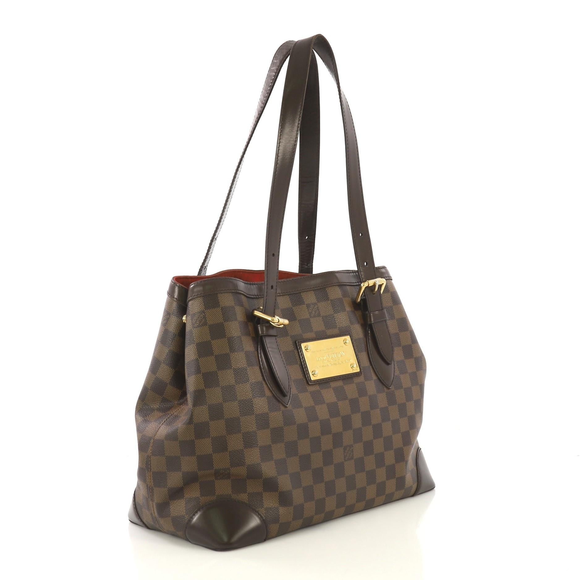 This Louis Vuitton Hampstead Handbag Damier MM, crafted from damier ebene coated canvas, features dual flat handles, leather trim, side snap closures, metal logo plate, and gold-tone hardware. Its clasp closure opens to a red microfiber interior