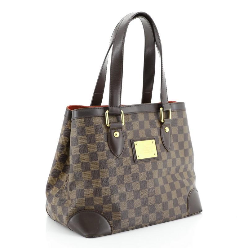 This Louis Vuitton Hampstead Handbag Damier PM, crafted from damier ebene coated canvas, features dual flat handles, leather trim, side snap closures, metal logo plate, and gold-tone hardware. Its clasp closure opens to a red microfiber interior