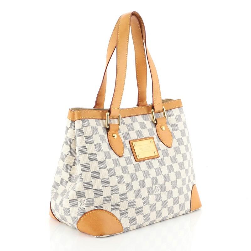 This Louis Vuitton Hampstead Handbag Damier PM, crafted from damier azur coated canvas, features dual flat handles, side snap closures, metal logo plate, and gold-tone hardware. Its clasp closure opens to a neutral microfiber interior with zip and