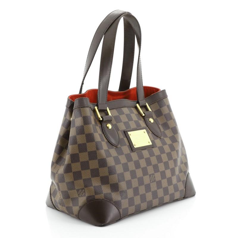 This Louis Vuitton Hampstead Handbag Damier PM, crafted from damier ebene coated canvas, features dual flat handles, leather trim, side snap closures, metal logo plate, and gold-tone hardware. Its clasp closure opens to a red microfiber interior