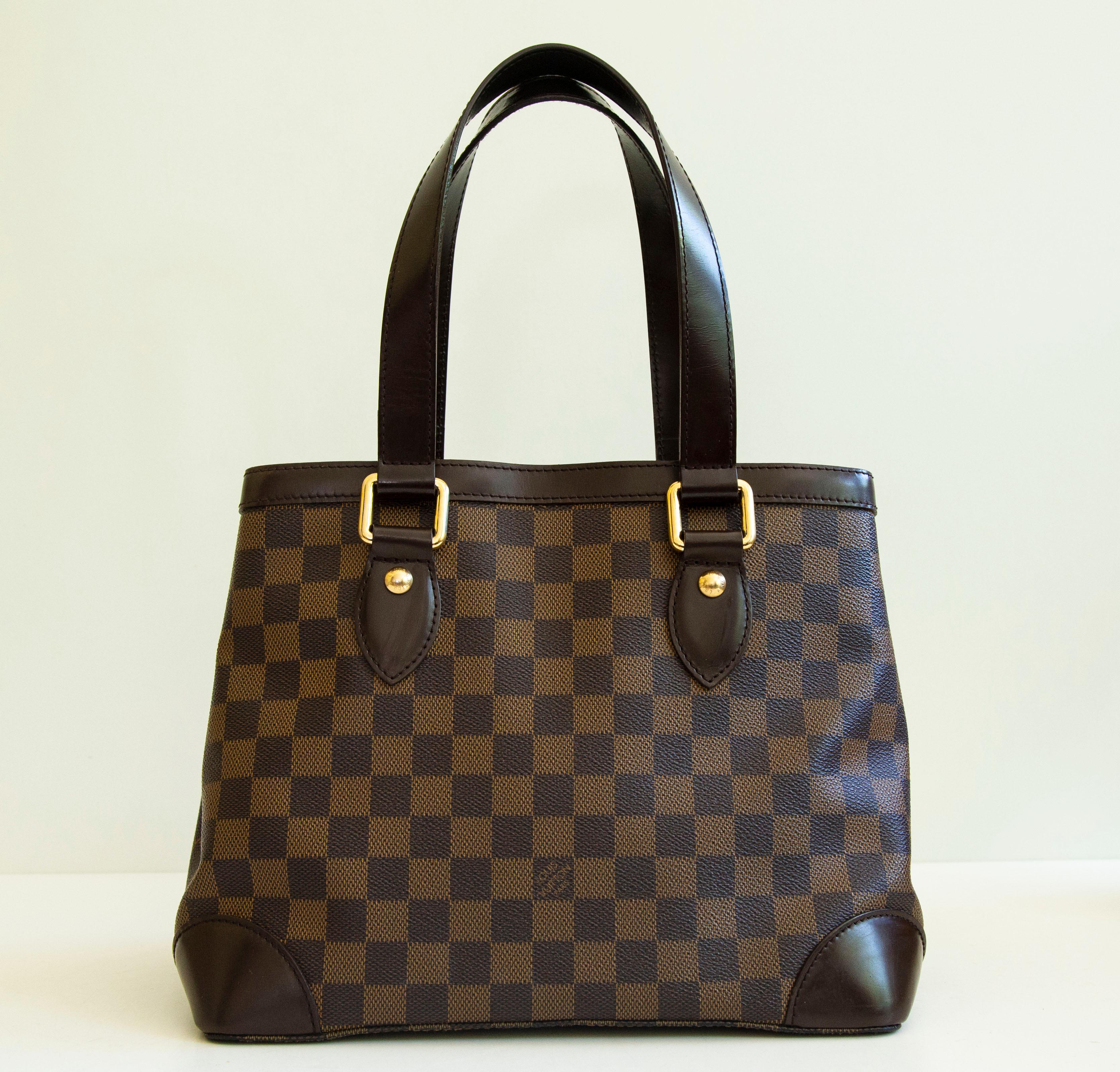 An authentic Louis Vuitton Hampstead PM. The bag features an Ebene Damier exterior, brown leather trim, and gold-toned hardware. The interior is lined with red fabric and there are three side pockets, one zipped pocket, and two slip pockets. The