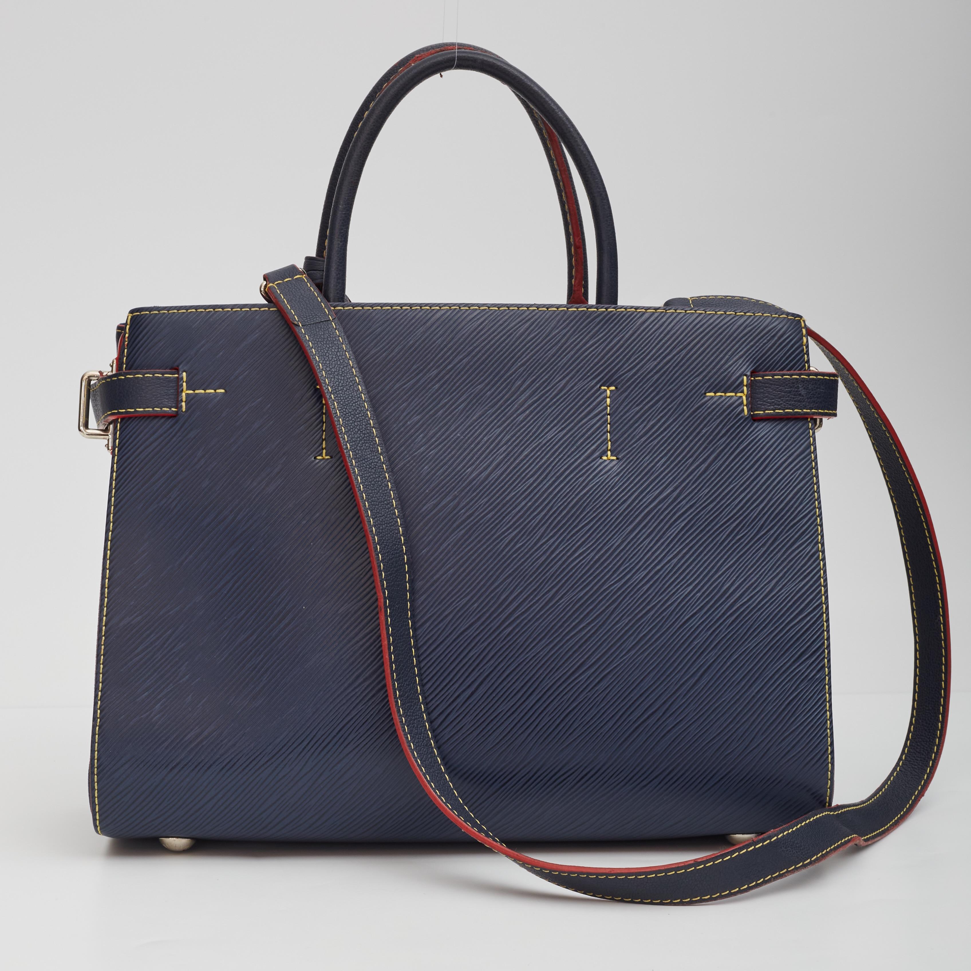 This tote is crafted of textured epi leather in dark blue. The handbag features red glazing trim, contrasting yellow stitching, adjustable leather side straps, and polished silver hardware including a frontal LV turn lock. Dual top handles and an