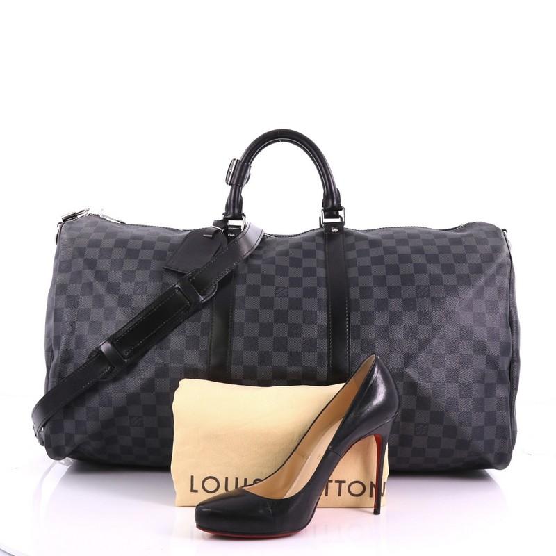 This Louis Vuitton Keepall Bandouliere Bag Damier Graphite 55, crafted with damier graphite coated canvas, features dual rolled handles, black leather trims, and silver-tone hardware. Its top zip closure opens to a navy fabric interior .