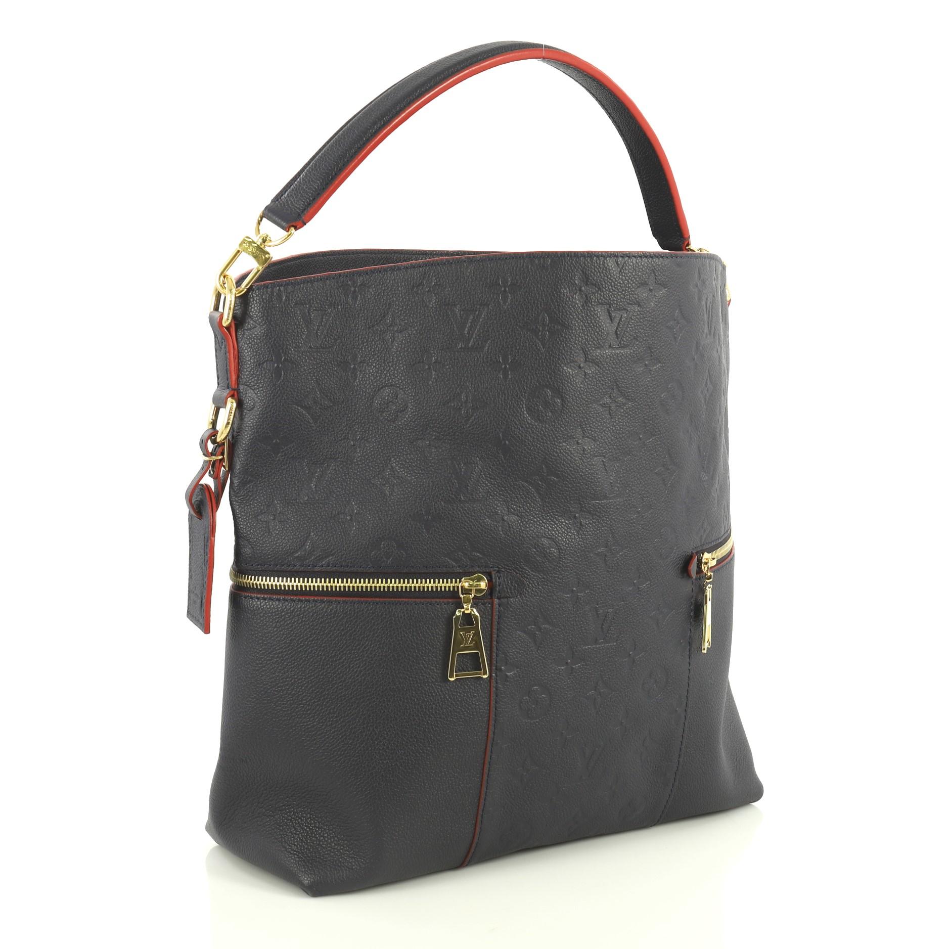 This Louis Vuitton Melie Handbag Monogram Empreinte Leather, crafted in blue monogram empreinte leather, features a single looped leather handle, two exterior side zip pockets, and gold-tone hardware. Its wide top opening displays a red fabric