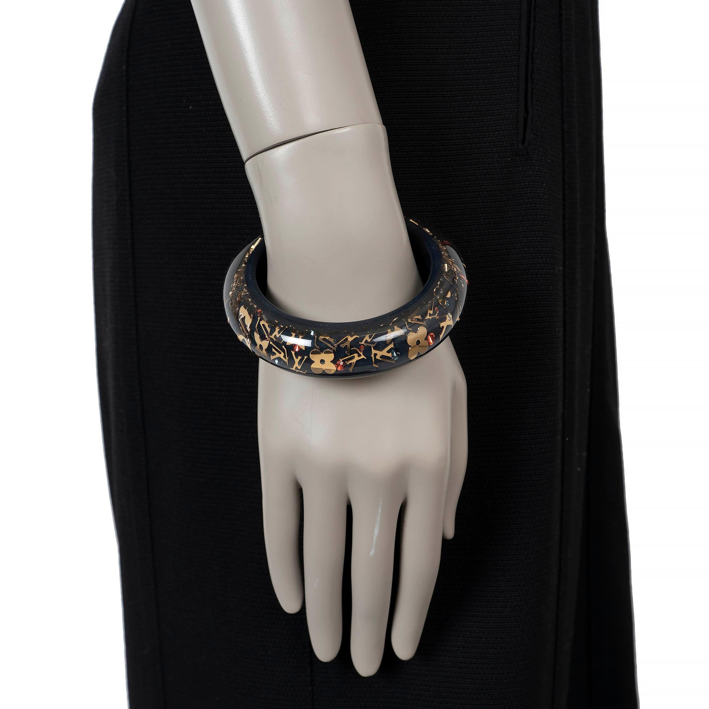 This 100% authentic Louis Vuitton Inclusion bangle bracelet is crafted from clear resin infused with gold-tone Monogram motifs and navy blue background, creating a striking play of depth and light. The wide silhouette makes it a bold statement