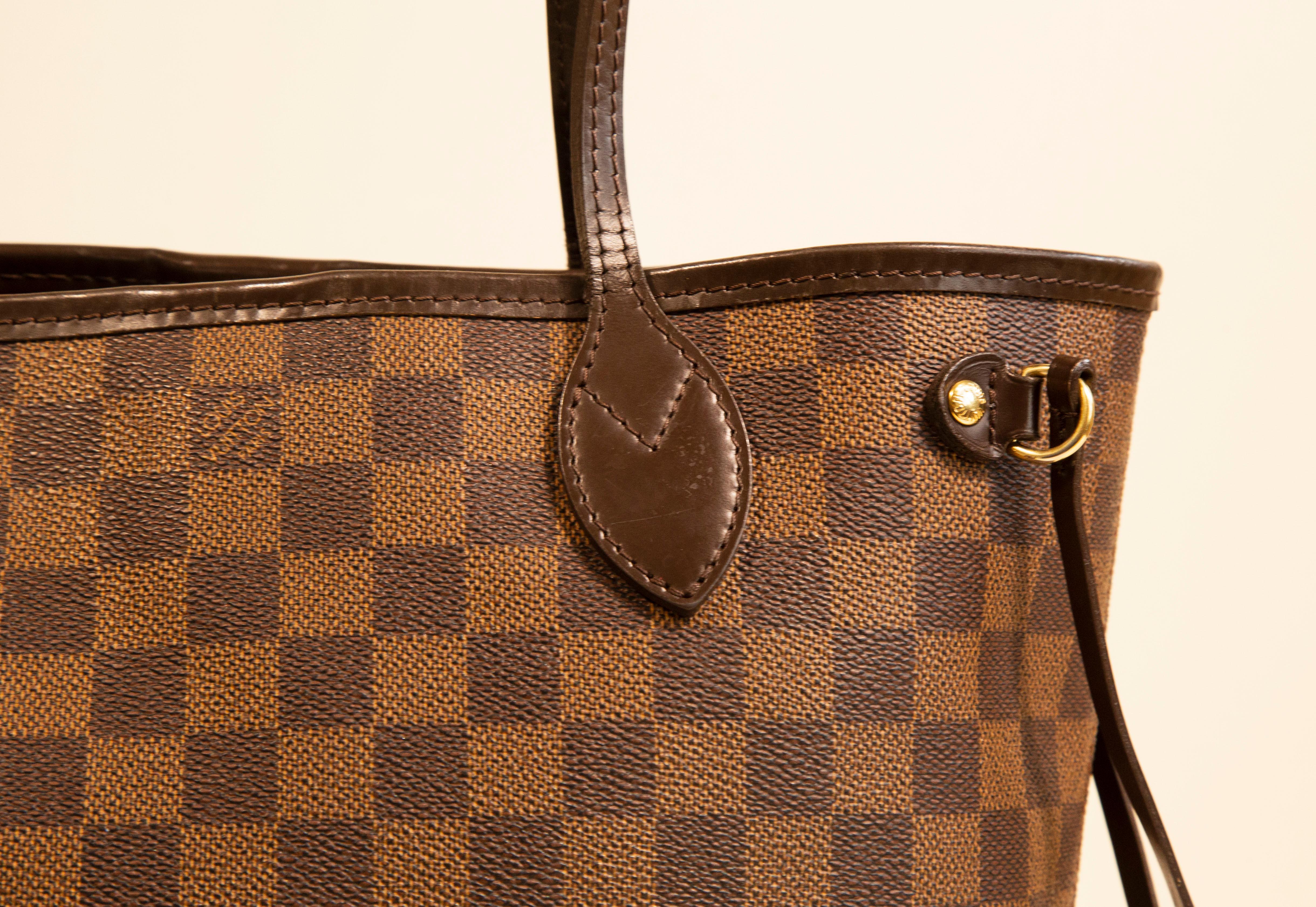 A Louis Vuitton Neverfull PM tote. The bag features a Damier Ebene exterior, with brown leather trim and gold-tone hardware. The interior is lined with red fabric. The exterior shows minor signs of use. The interior is clean, but there a few small