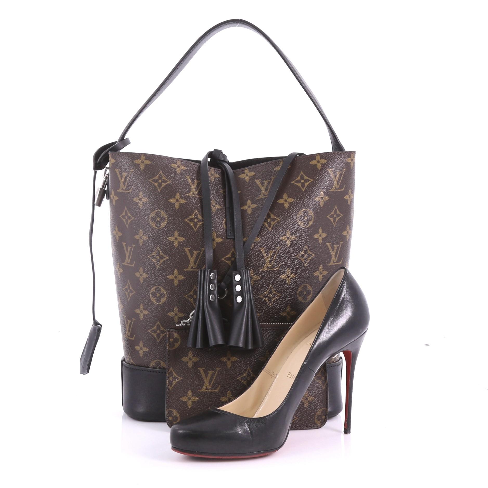 This Louis Vuitton NN14 Idole Bucket Bag Monogram Canvas and Leather GM, crafted from brown monogram coated canvas and black leather, features hand-pleated leather tassels, flat handle, and silver-tone hardware. It opens to a black microfiber