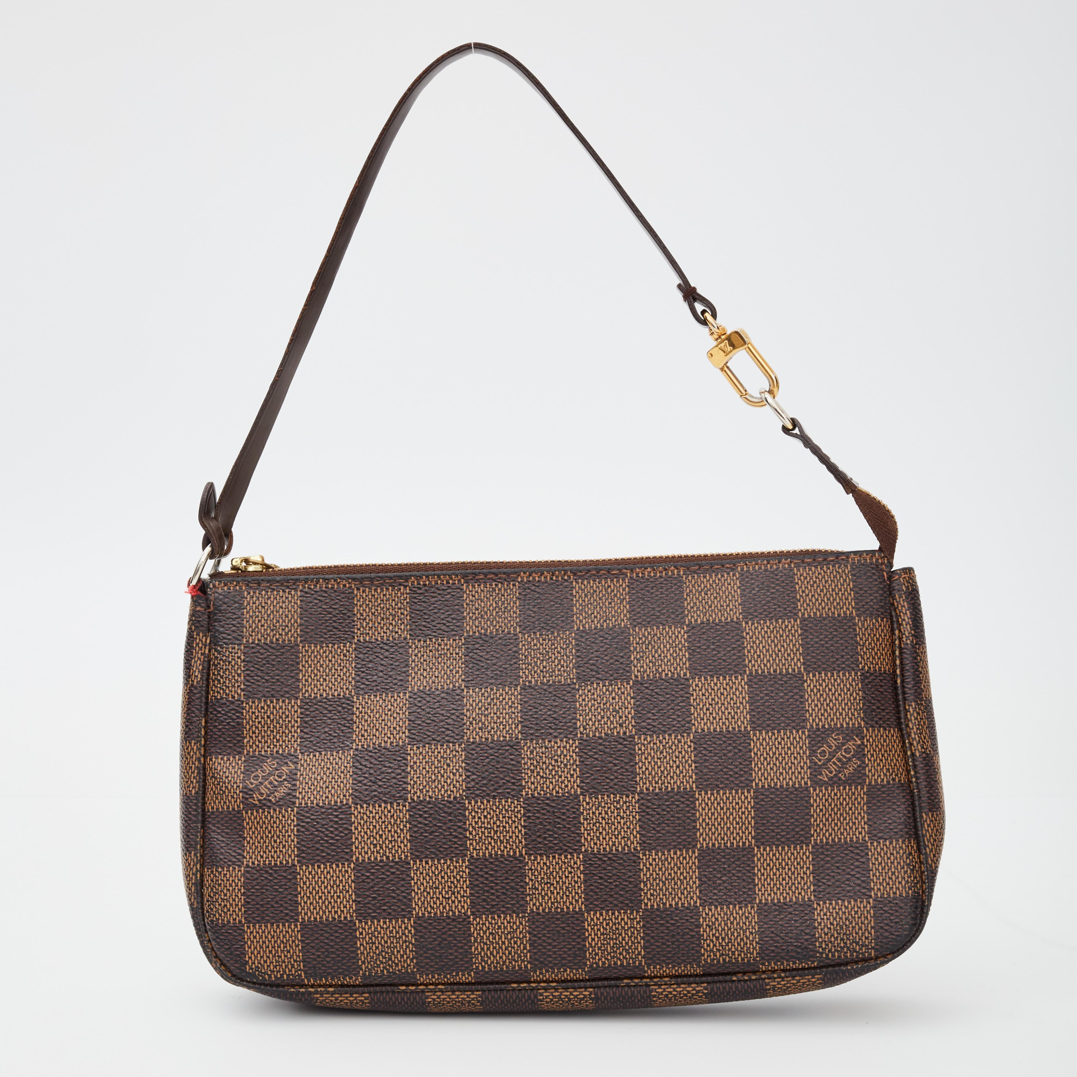 This little pochette bag is made with damier ebene coated canvas and features treated leather details including a removable flat leather top strap, top zip closure, gold tone hardware and red woven fabric interior lining.

COLOR: Brown
MATERIAL: