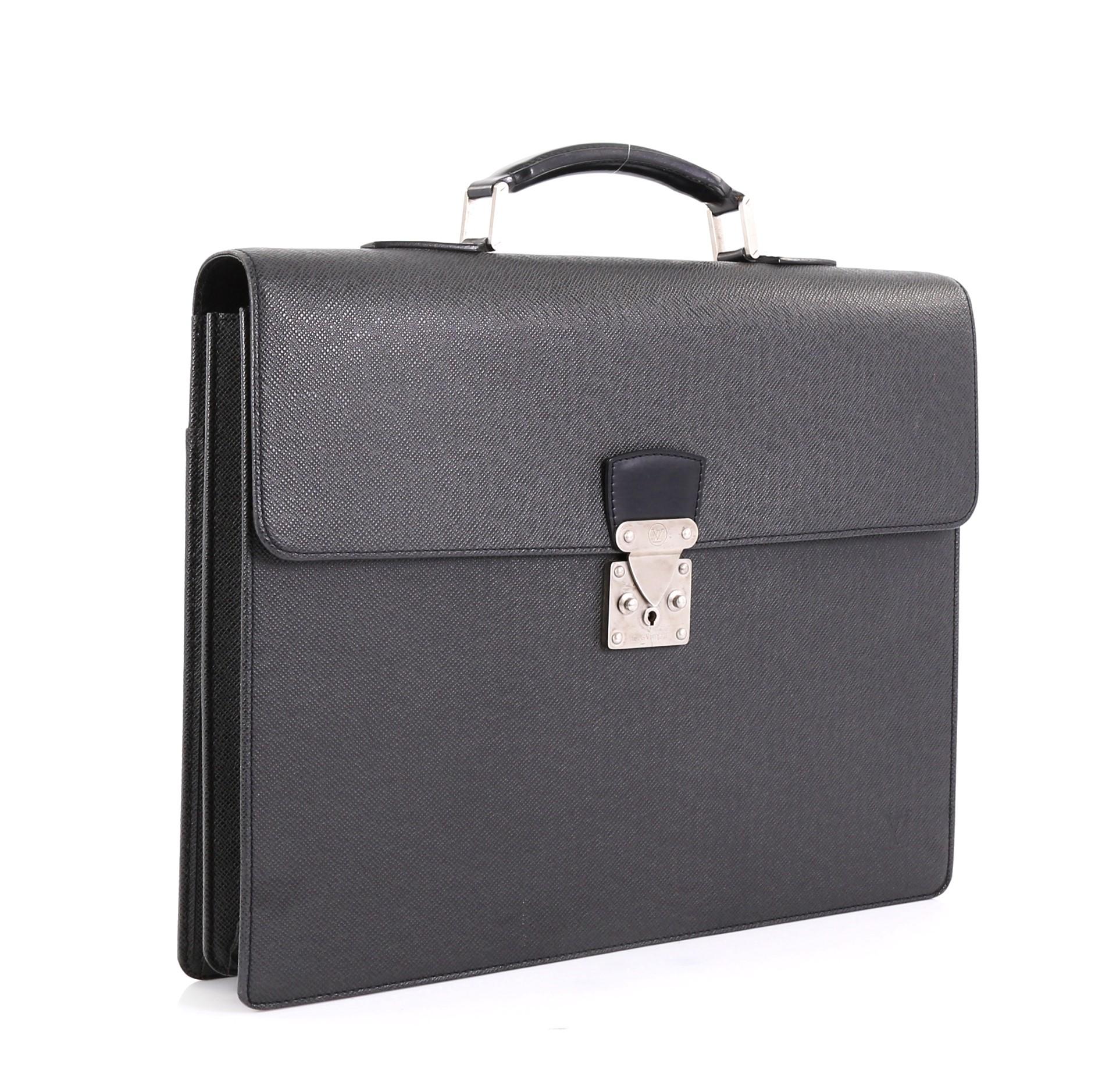 This Louis Vuitton Serviette Moskova Briefcase Taiga Leather, crafted in black taiga leather, features a flat top handle, exterior back flat pocket, and silver-tone hardware. Its S-lock closure opens to a black leather interior divided into two