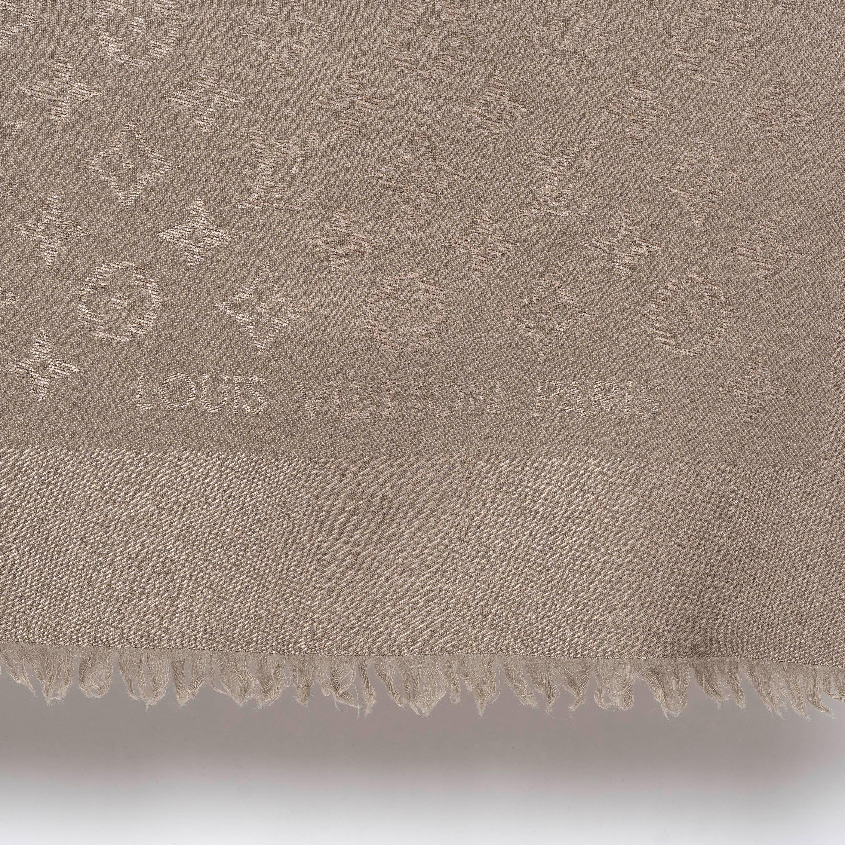 This 100% authentic Louis Vuitton Monogram Classic shawl in taupe is crafted from a luxurious blend of silk (60%) and wool (40%). It features a jacquard weave that gives the tone-on-tone fabric a subtle sheen, complemented by fringed edges for a