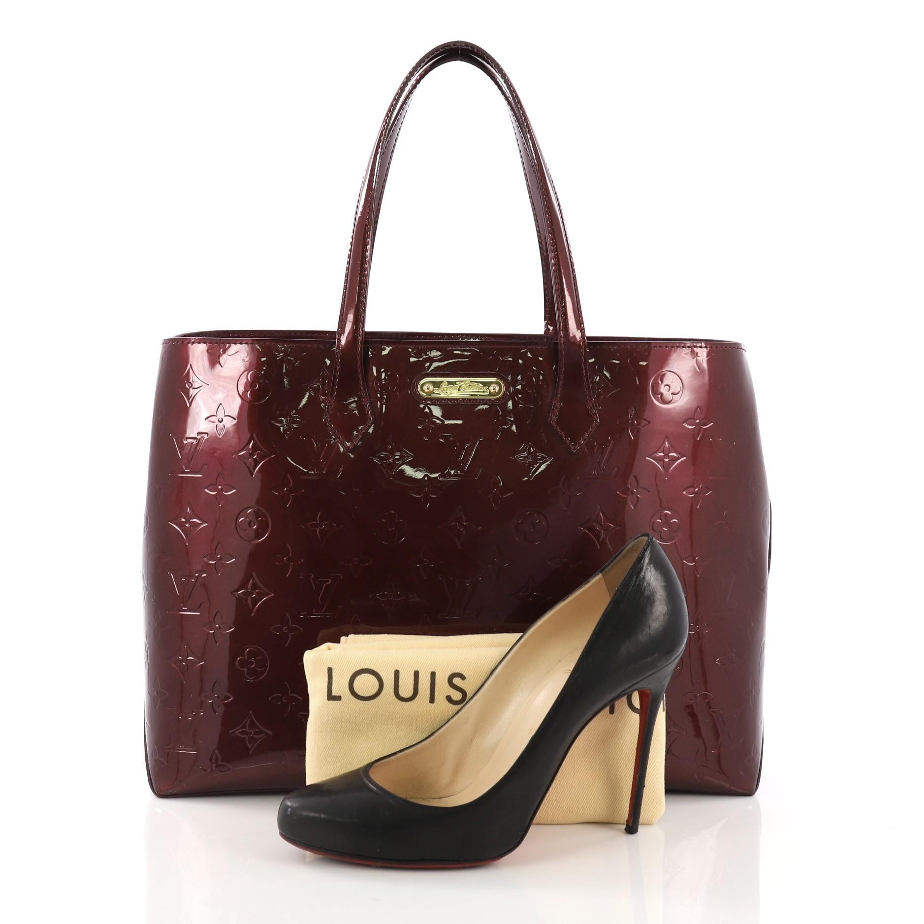 This Louis Vuitton Wilshire Handbag Monogram Vernis MM, crafted in burgundy monogram vernis leather, features dual flat handles and gold-tone hardware. Its wide top with hook closure opens to a burgundy fabric interior with zip and slip pockets.