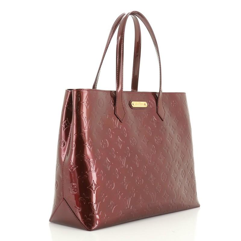 This Louis Vuitton Wilshire Handbag Monogram Vernis MM, crafted in red monogram vernis leather, features dual flat handles and gold-tone hardware. Its wide top with hook closure opens to a red fabric interior with zip and slip pockets. Authenticity