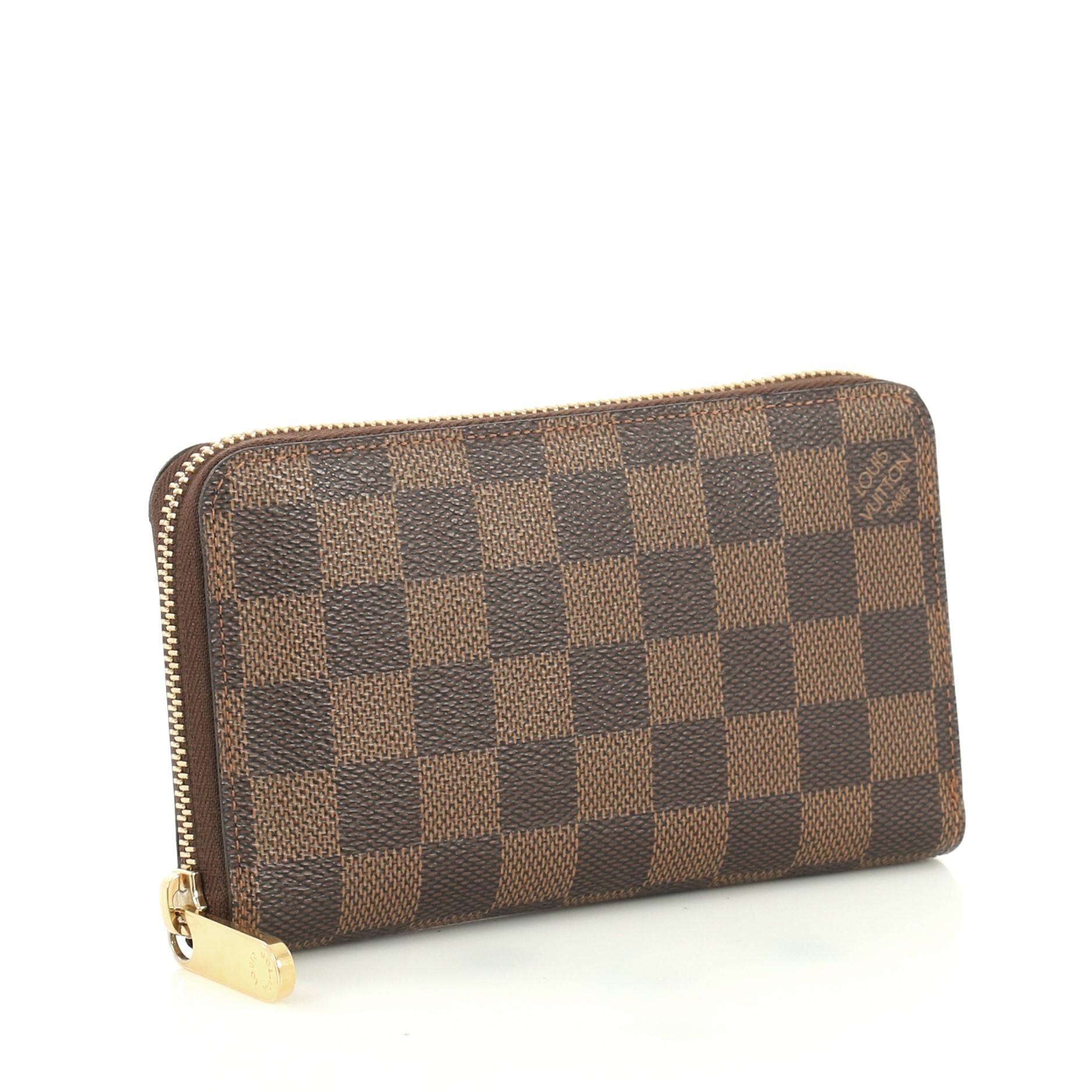 This Louis Vuitton Zippy Compact Wallet Damier, crafted from damier ebene coated canvas, features gold-tone hardware. Its zip-around closure opens to a brown leather interior with multiple card slots,zip pocket, and slip pockets. Authenticity code