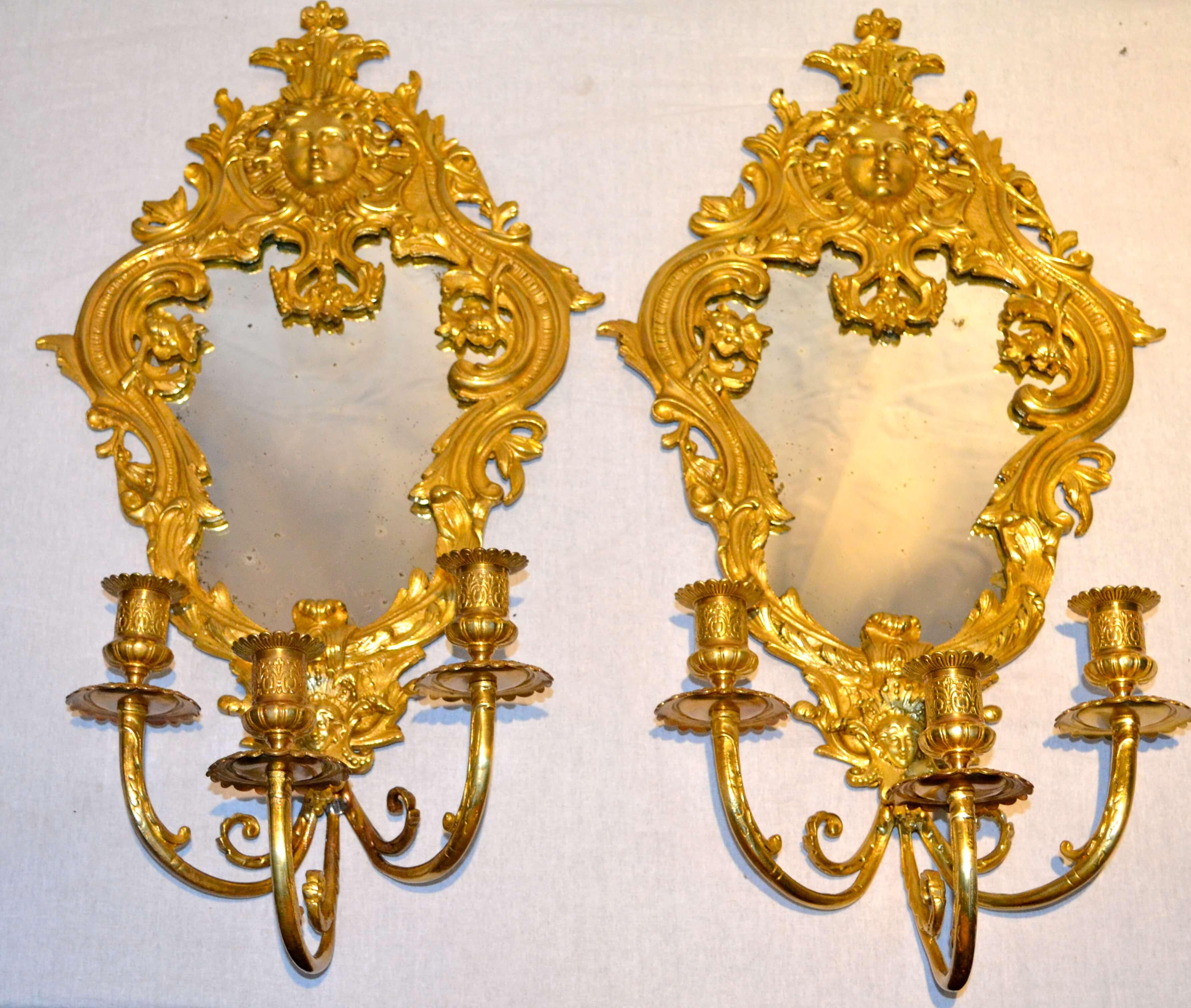 A pair of Louis XIV-style mirrored brass sconces with three candle holders, the mirror framed in scrolling rococo foliate trim with classical masks at the bottom and top of each sconce. Unwired.