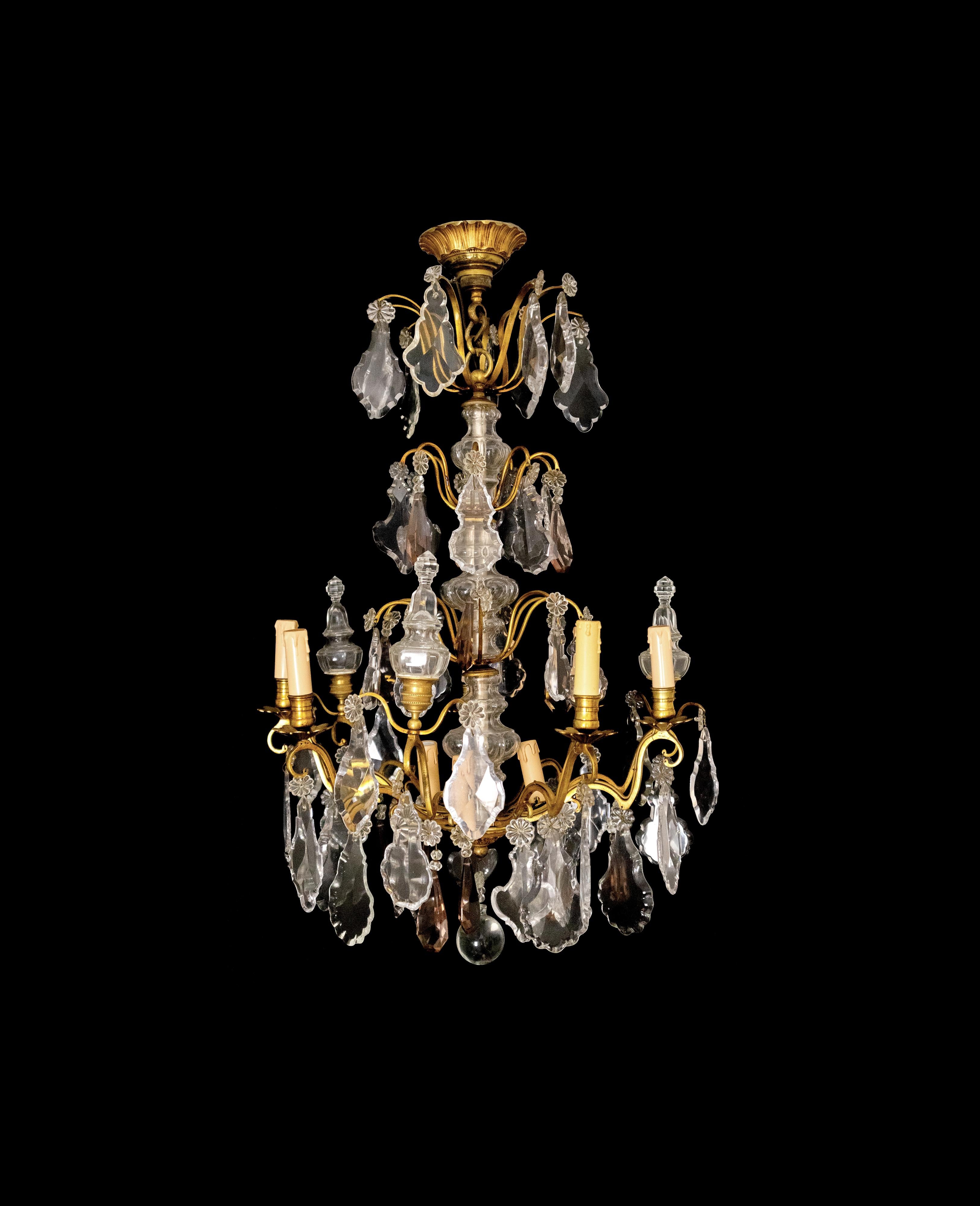 This Louis XV-Style bronze chandelier from the 19th Century features a refined design with crystal pendants and finials in tinted glass. 
Equipped with nine light sockets and candlestick holders. 
The combination of gold-toned bronze and hand-cut
