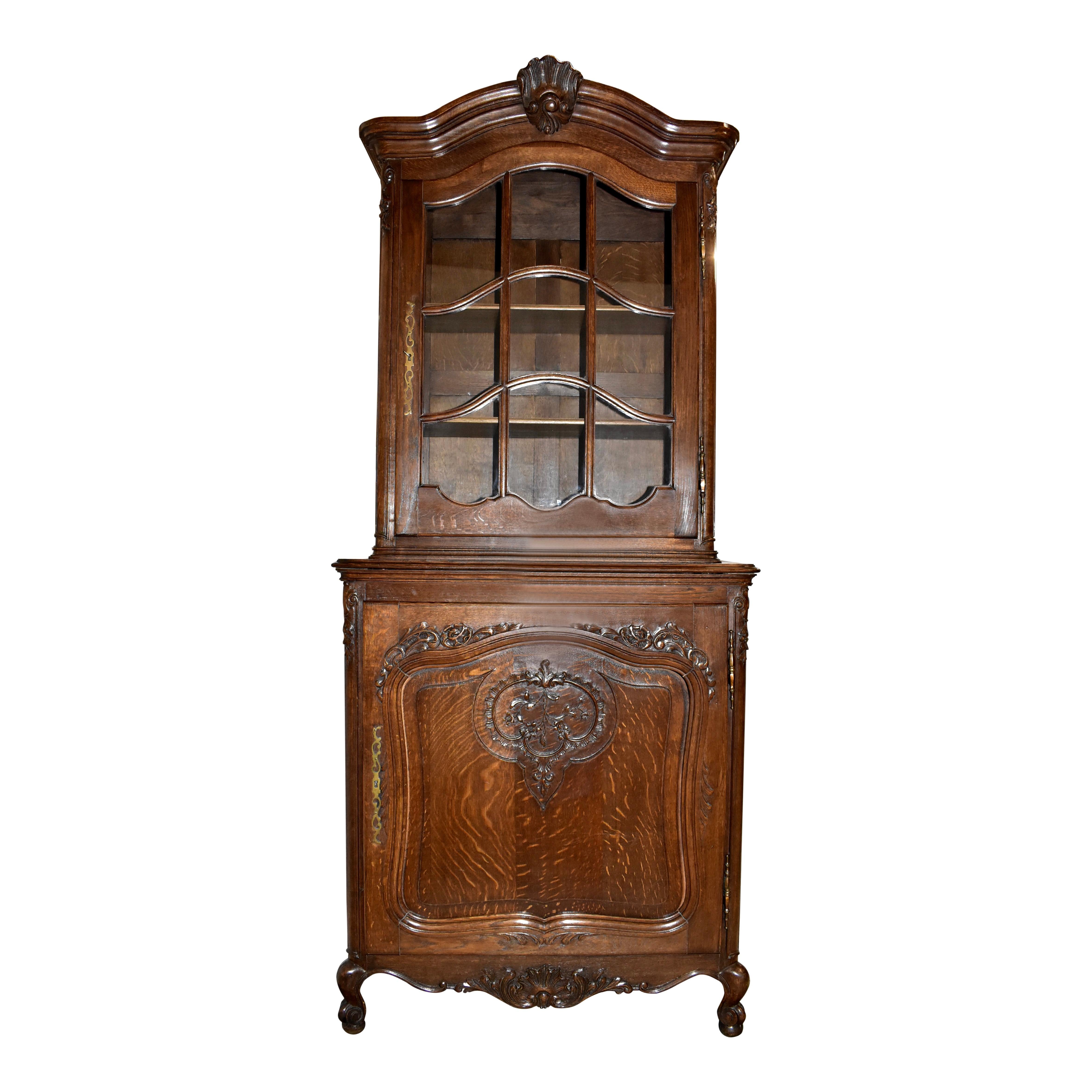 This elegant double bodied vitrine pairs solid oak construction with the soft, graceful lines of an arched pediment, rounded front corners, and cabriole legs. The top portion showcases an arched crown with a central carved shell over an arched glass