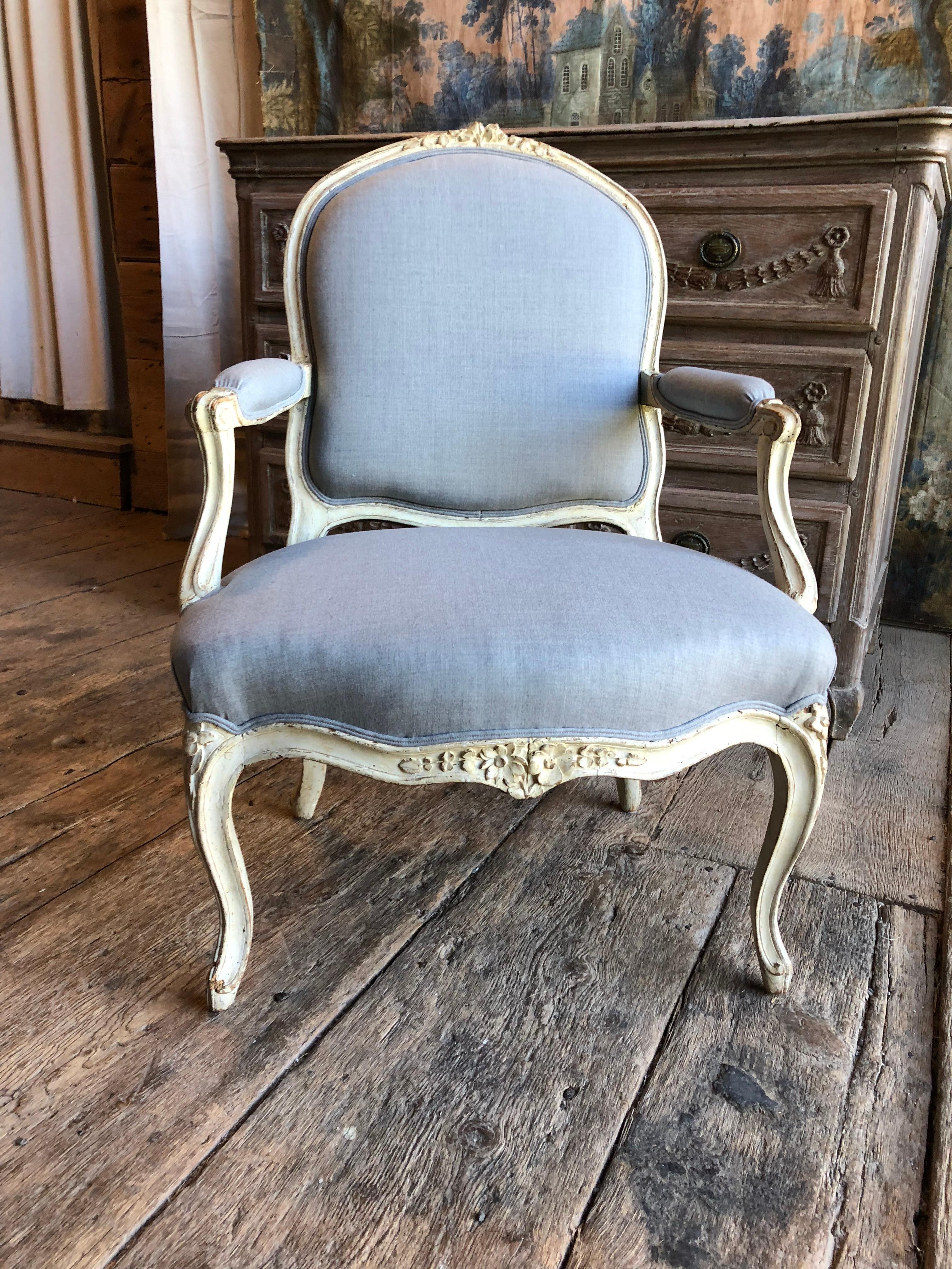 A charming Louis XV period fauteuil cabriolet in its original painted finish, with floral carvings on the back and seat rail, cabriole legs and a scalloped apron. Recently upholstered in grey/blue linen fabric. French, circa 1760.