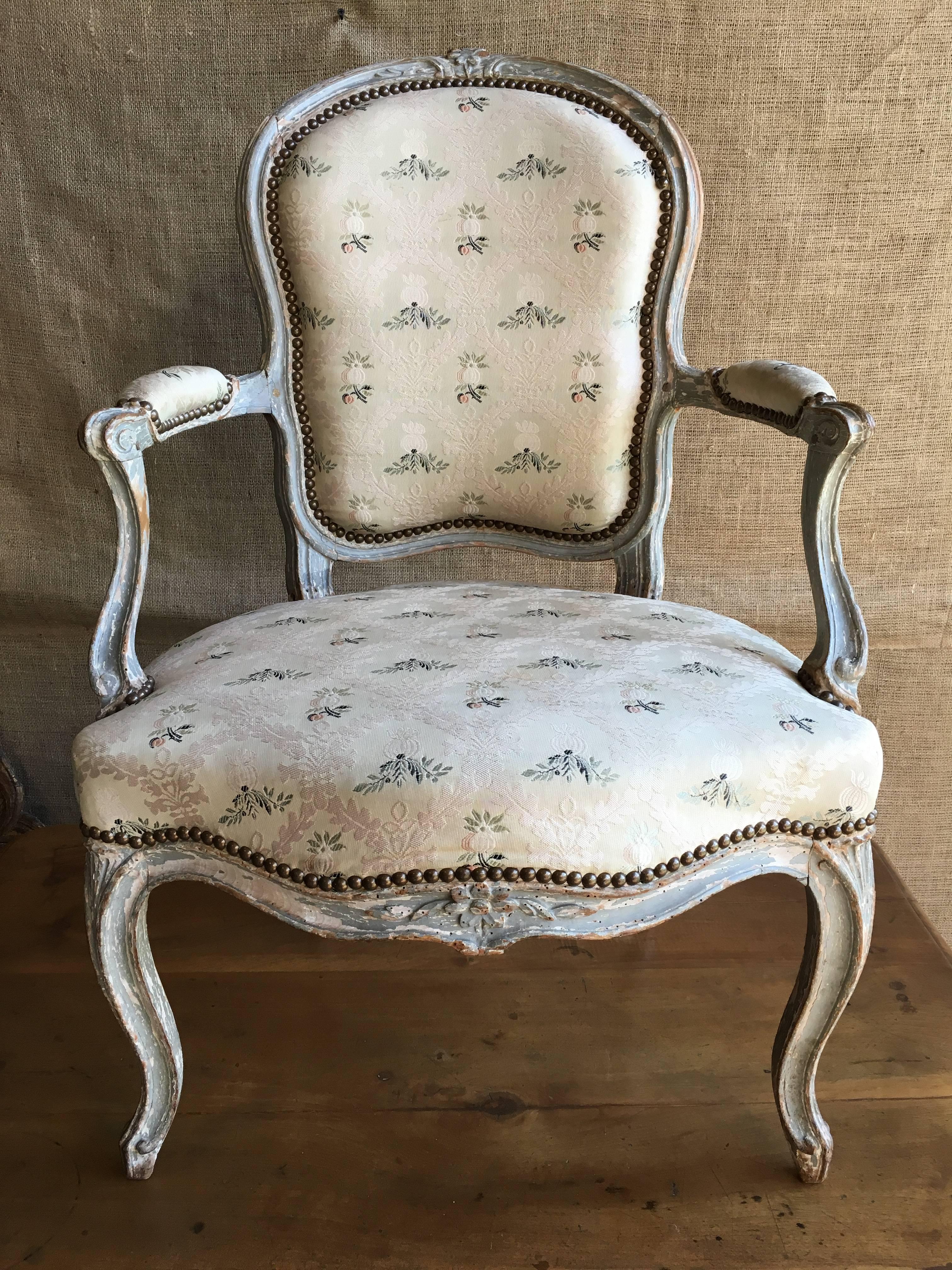 A lovely French Louis XV period armchair in beech retaining its original grey painted finish and currently upholstered in an antique brocade fabric, nicely carved with a crest of flowers on the top rail, and duplicated on the seat rail, the chair is