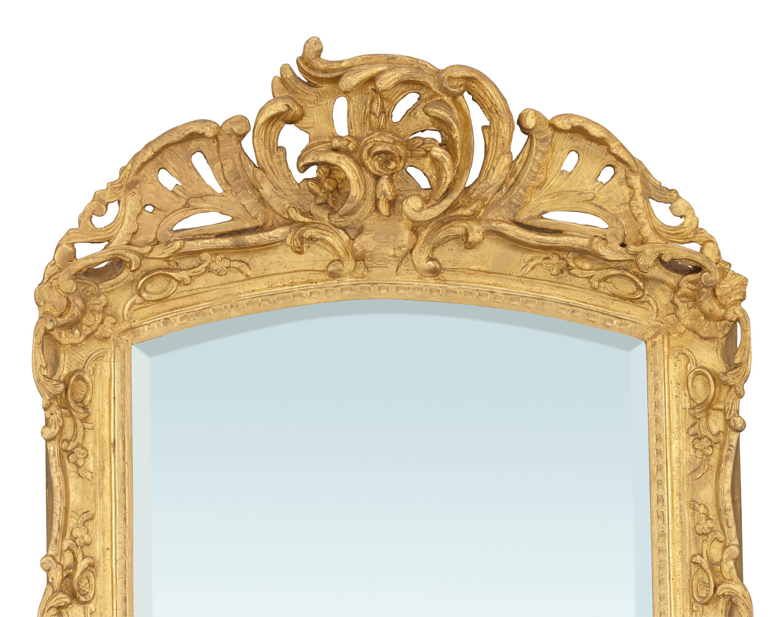 Commanding in size and artistry, this rare Louis XV-period gilt mirror is a tour-de-force of the era's highly ornamental, yet elegant style. The golden frame features sweeping, heavy scrolls and acanthus with floral and shell details that surround