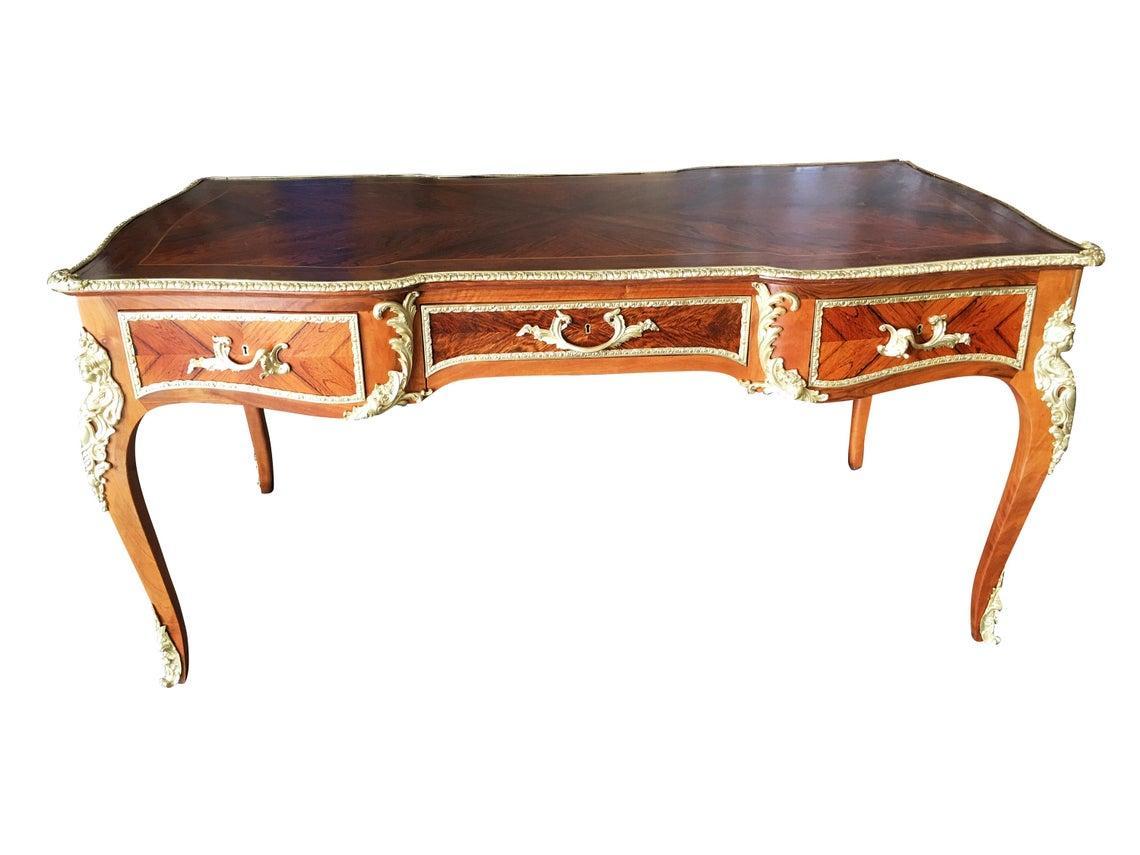 Vintage midcentury era Louis XV Rococo style executive writing desk made from mahogany, satinwood, and bureau Plat with hand-cast bronze accents. It features three lockable horizontal frieze drawers in an apron with foliate bronze trim, the reverse