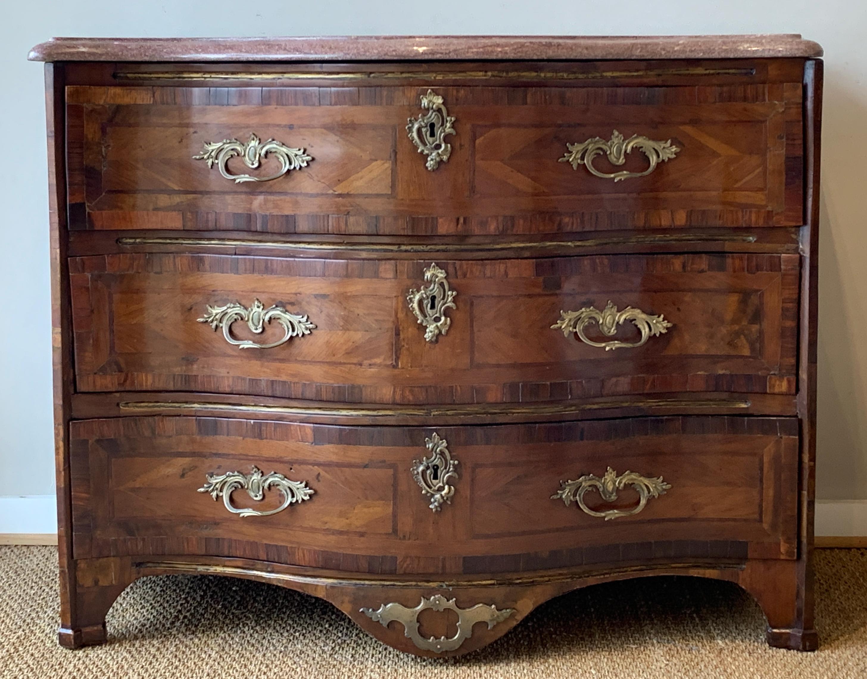 An elegant mid-18th century Louis XV serpentine front three drawer commode with later rose colored granite top. The elaborately inlaid exotic wood is accented with brass inlay between the drawers and appears to have the original cast brass pulls.