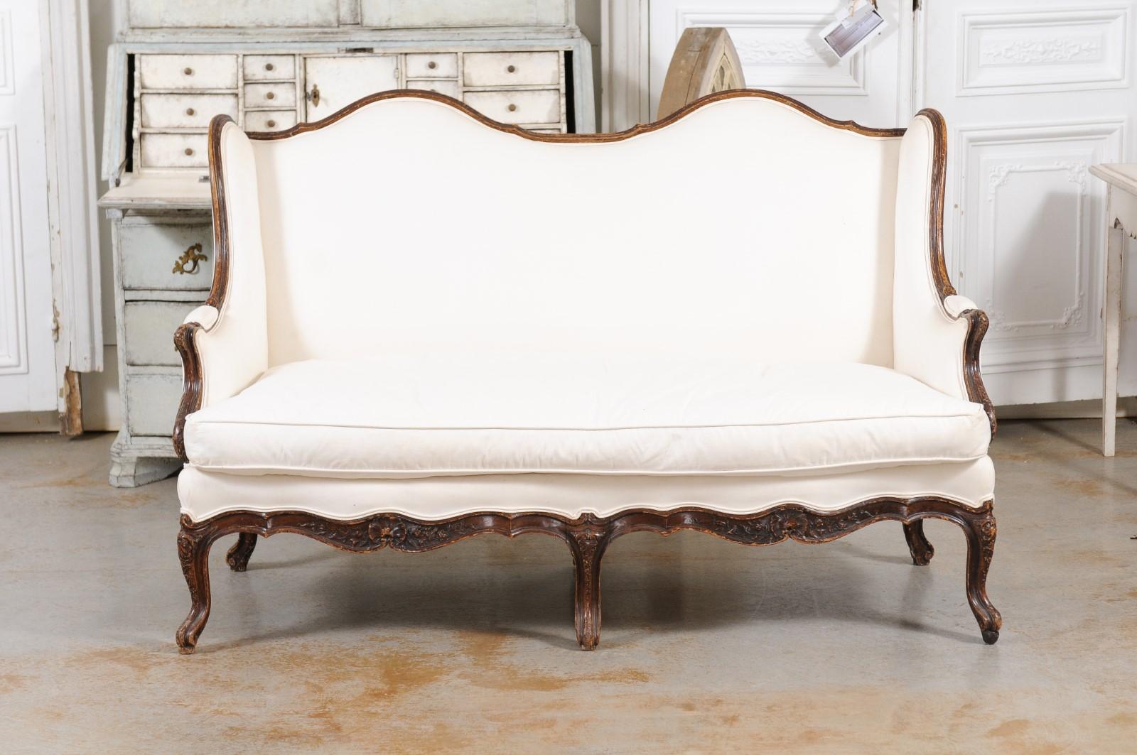 A French Louis XV style walnut canapé à oreilles from the late 19th century, with double camelback, cabriole legs and upholstery. Created in France during the later years of the 19th century, this two-seat canapé features a slightly slanted back