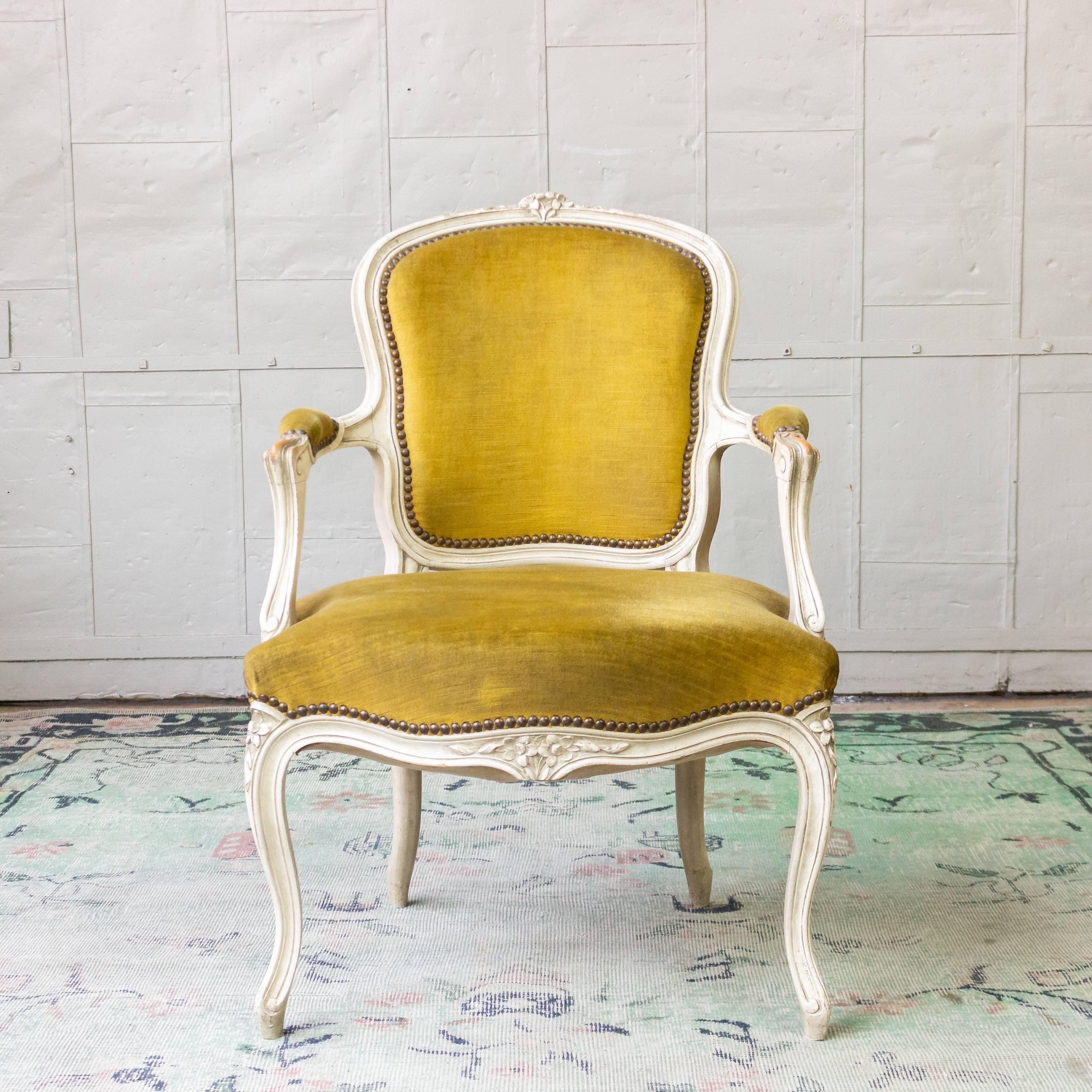 An elegant Louis XV armchair in gold velvet. Experience classic French luxury with this stunning vintage Louis XV armchair. With its white patinated finish, ornately carved wooden frame, padded arms, and decorative nailheads, this strong statement