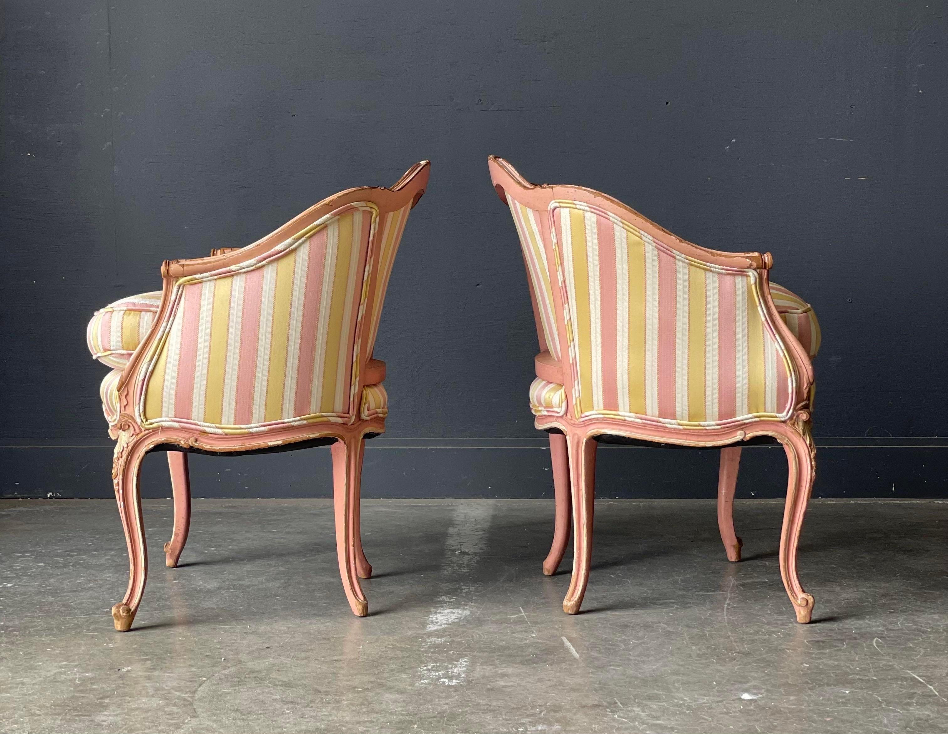 French Louis XV style bergere armchairs, perfect for your salle de séjour. Walnut frames with a distressed pink and white finish are upholstered in a gold, pink & white striped silk blend fabric. Features carvings to crest rail, arms, apron and