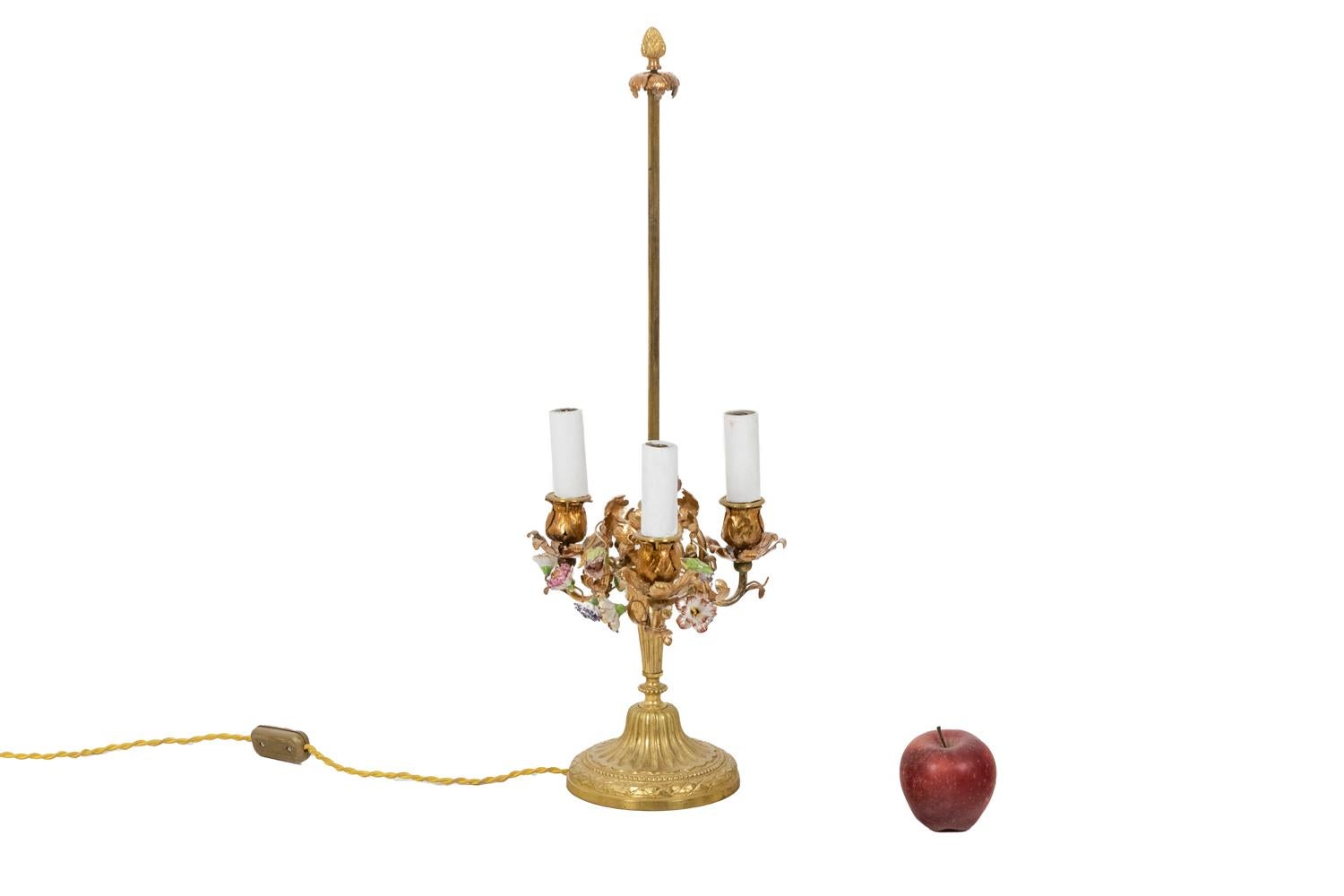 Bouillotte lamp in Louis XV style, in gilded brass decorated with porcelain flowers in pink, yellow and blue tones, with three lights. Round base, decorated with gadroons and a beaded frieze, fluted shaft and stem ending and top part with a stylized