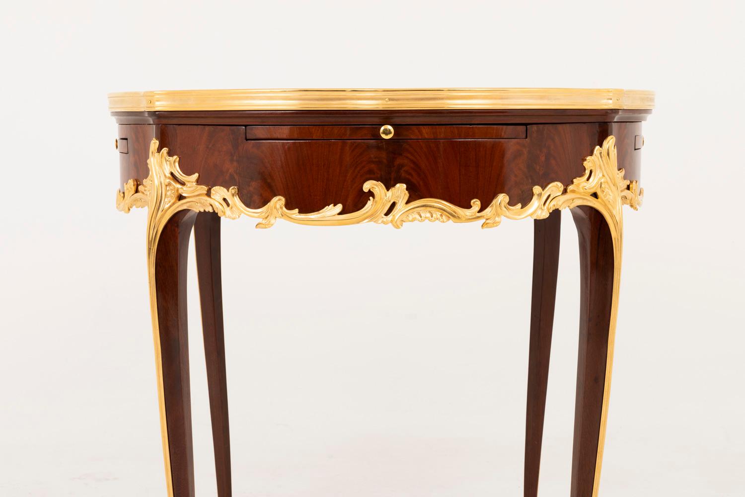 Table bouillotte circulaire de style Louis XV en marqueterie de bois de rose reposant sur quatre pieds cabriole.
Quatre feuilles de tablier à bord frontal incurvé recouvert de cuir brun frappé de fers dorés à décor de rinceaux feuillagés et ouvrant