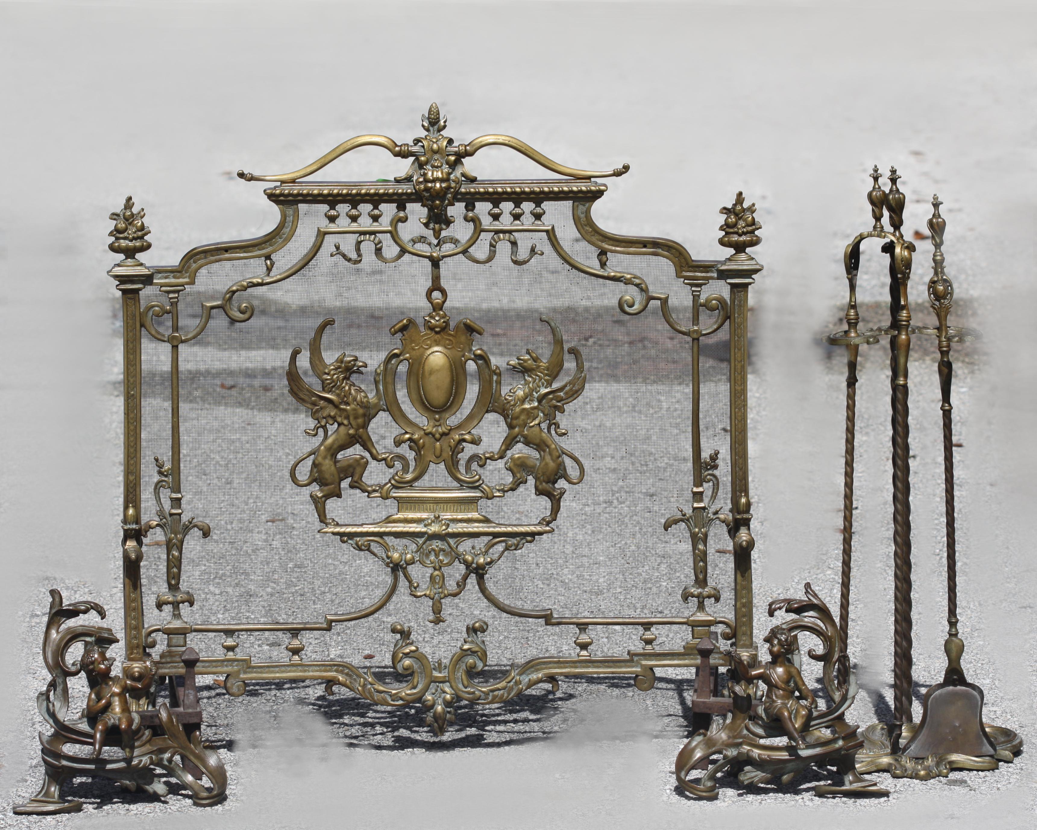 
Louis XV Style Bronze Fireplace Suite 
Late 19th Century. Comprising, a screen with serpentine cresting headed by a Baroque mask, a pair of winged griffins centering an armorial shield, a pair of leaf-scrolled chenets mounted with seated putti, and