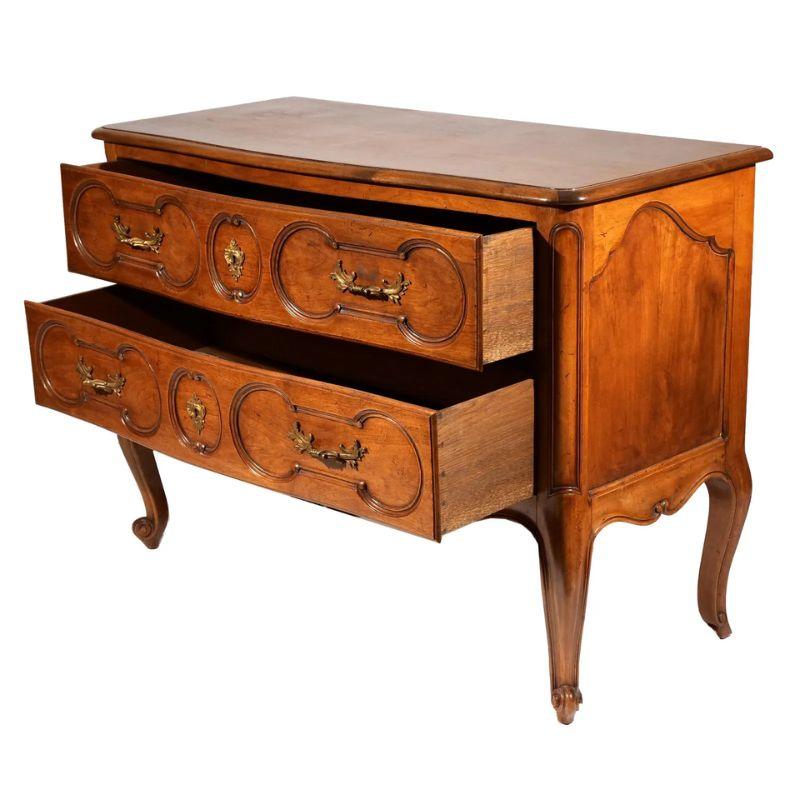 A Louis XV style carved commode with brass fittings. The two drawer wood chest embodies the essence of the Louis XV style, renowned for its lavish details and curvaceous aesthetics. Crafted with  attention to detail, it is an embodiment of the
