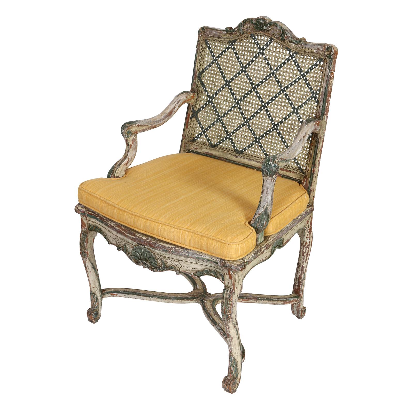 A Louis XV style painted fauteuil with a caned back and yellow seat cushion. The French armchair is painted with a blue trellis diamond grid to both sides of the back. Curved chair legs meet with an X stretcher. Chair back is square with an arched