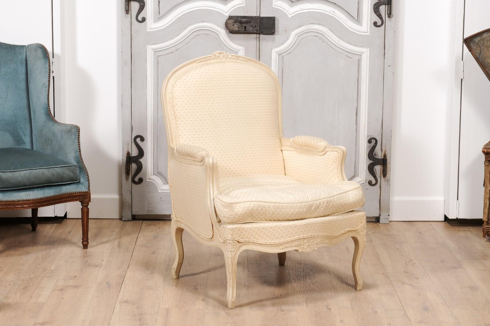A pair of French Louis XV style bergères chairs from the 20th century with cream painted finish, floral carved crest, knees and apron, and cabriole legs. Exuding timeless elegance, this pair of French Louis XV style bergères chairs from the 20th
