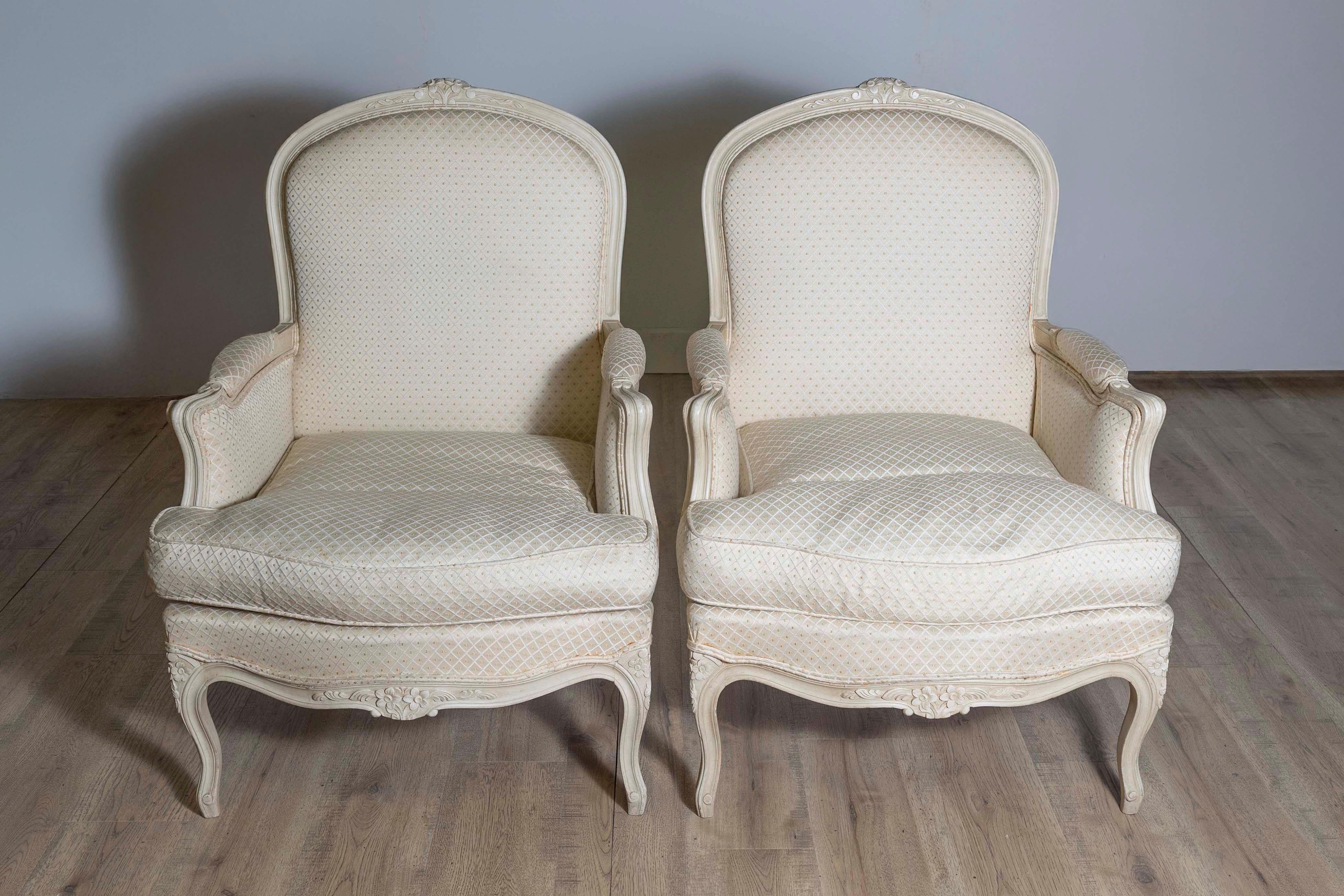 A pair of French Louis XV style bergères chairs from the 20th century with cream painted finish, floral carved crest, knees and apron, and cabriole legs. Exuding timeless elegance, this pair of French Louis XV style bergères chairs from the 20th